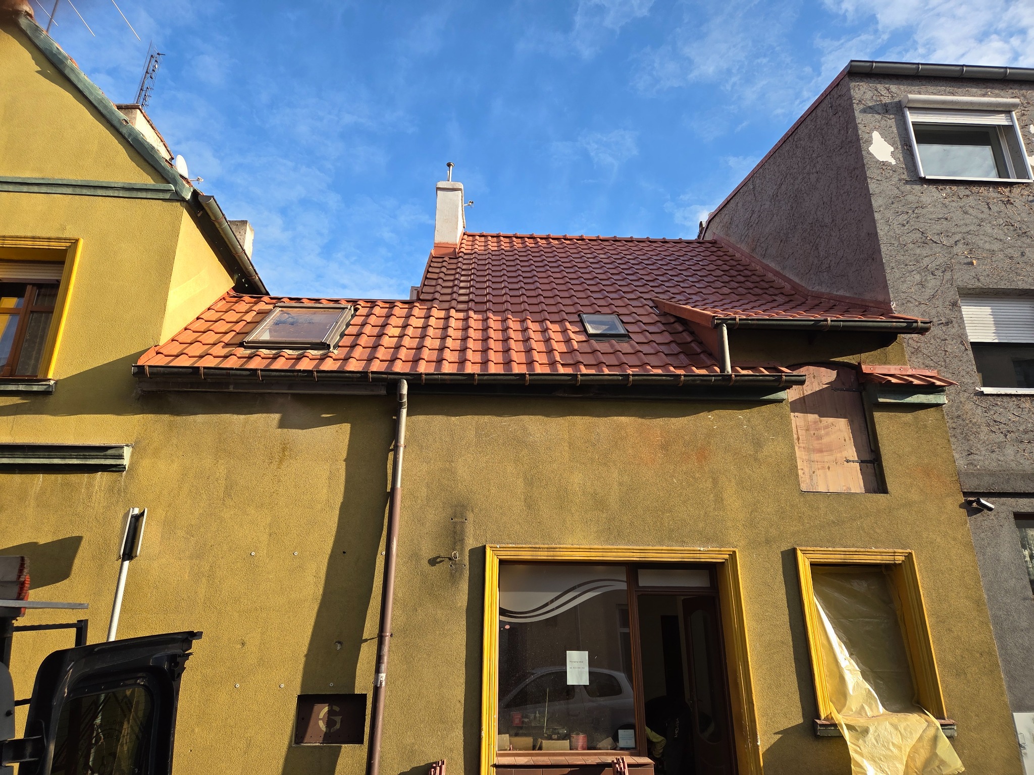 Czerwona dachówka na budynku z oknami dachowymi i kominem, widok z dołu na tle błękitnego nieba. Fragment samochodu w lewym dolnym rogu.