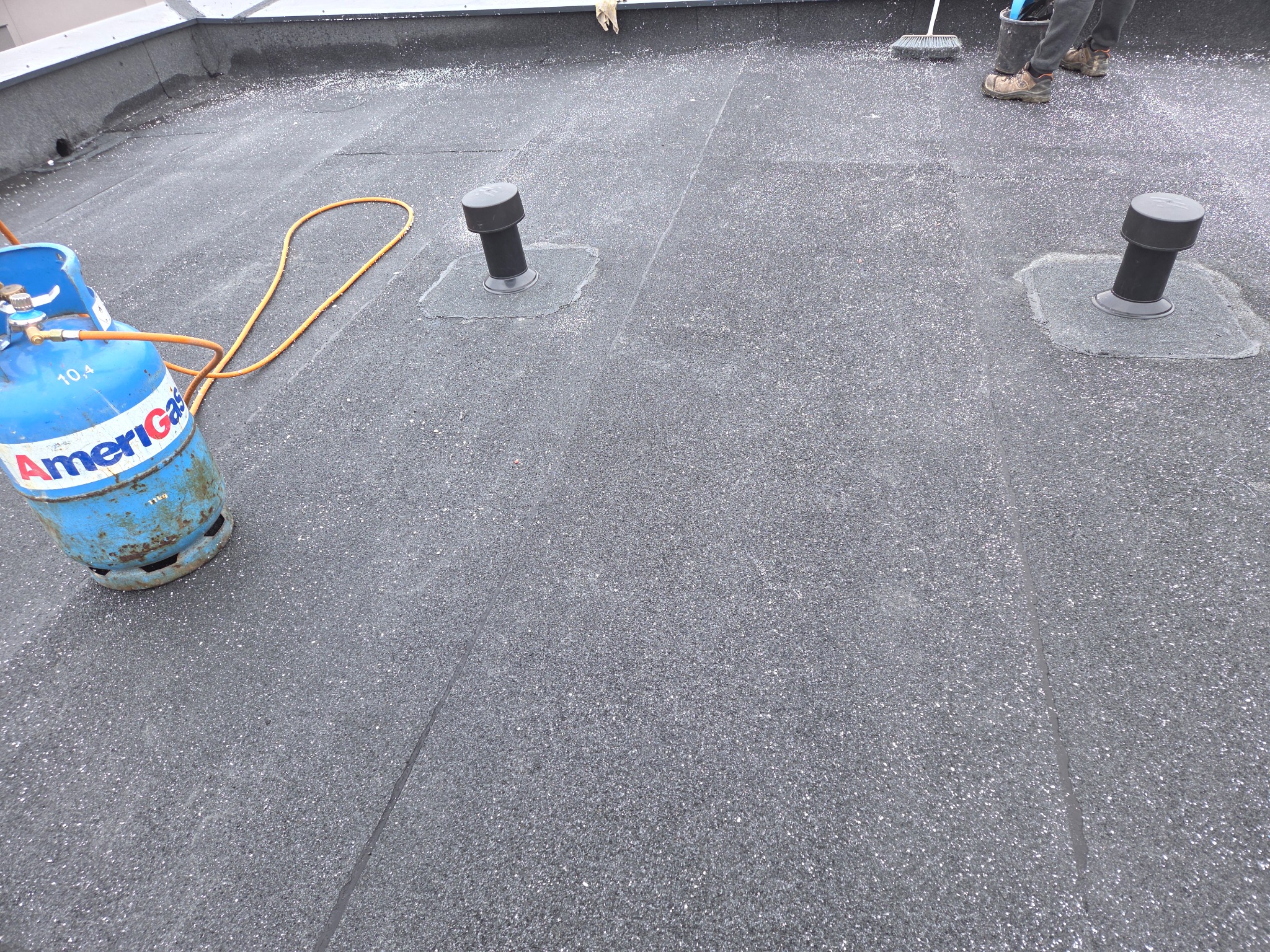 Płaski dach pokryty papą z widocznymi kominkami wentylacyjnymi i butlą gazową Amerigas. W tle osoba z miotłą. Powierzchnia dachu pokryta drobnym kruszywem.