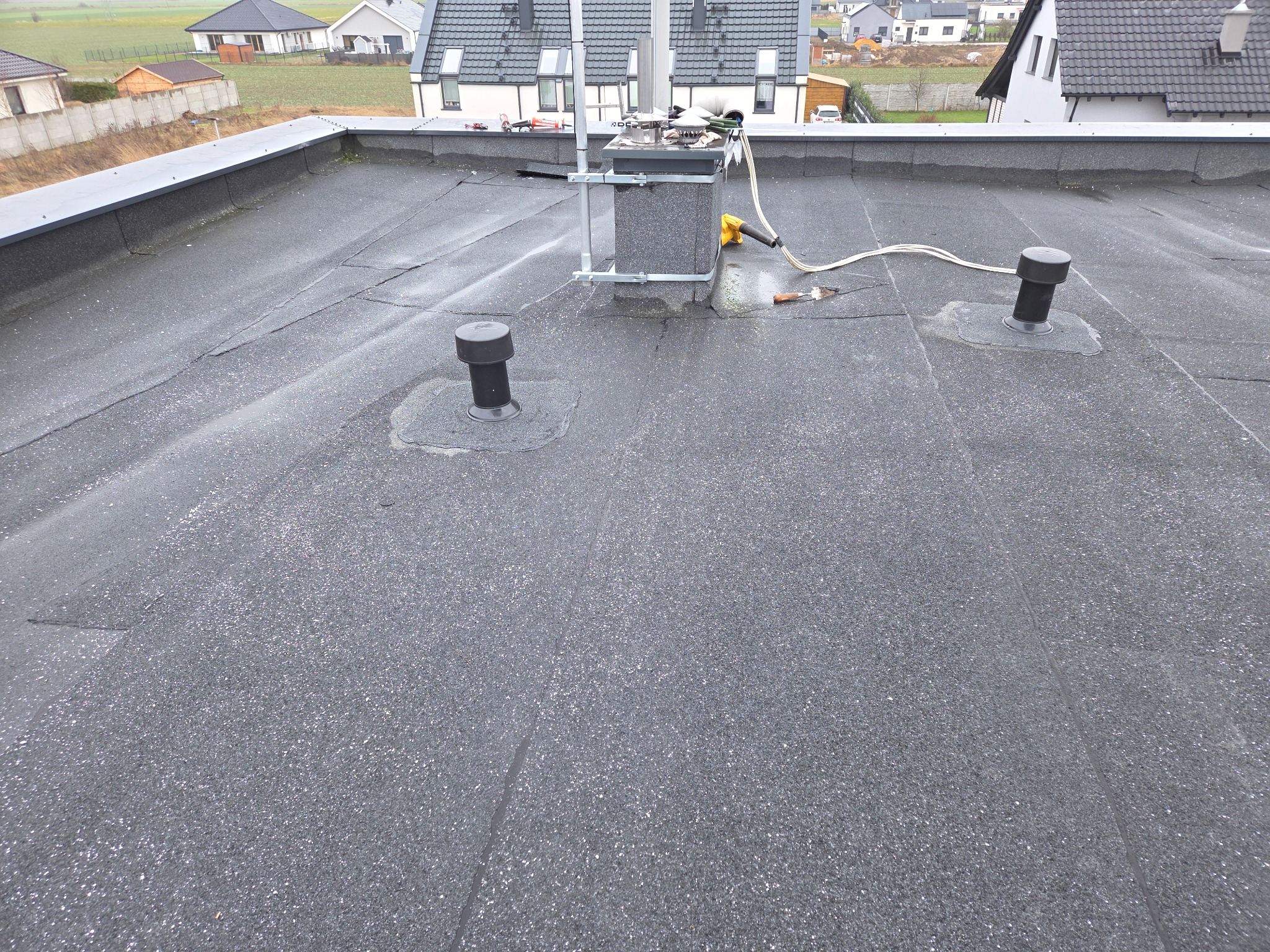 Płaski dach pokryty papą termozgrzewalną z kominem i odpowietrznikami. Widoczne narzędzia dekarskie. Tło stanowią domy jednorodzinne i pole.