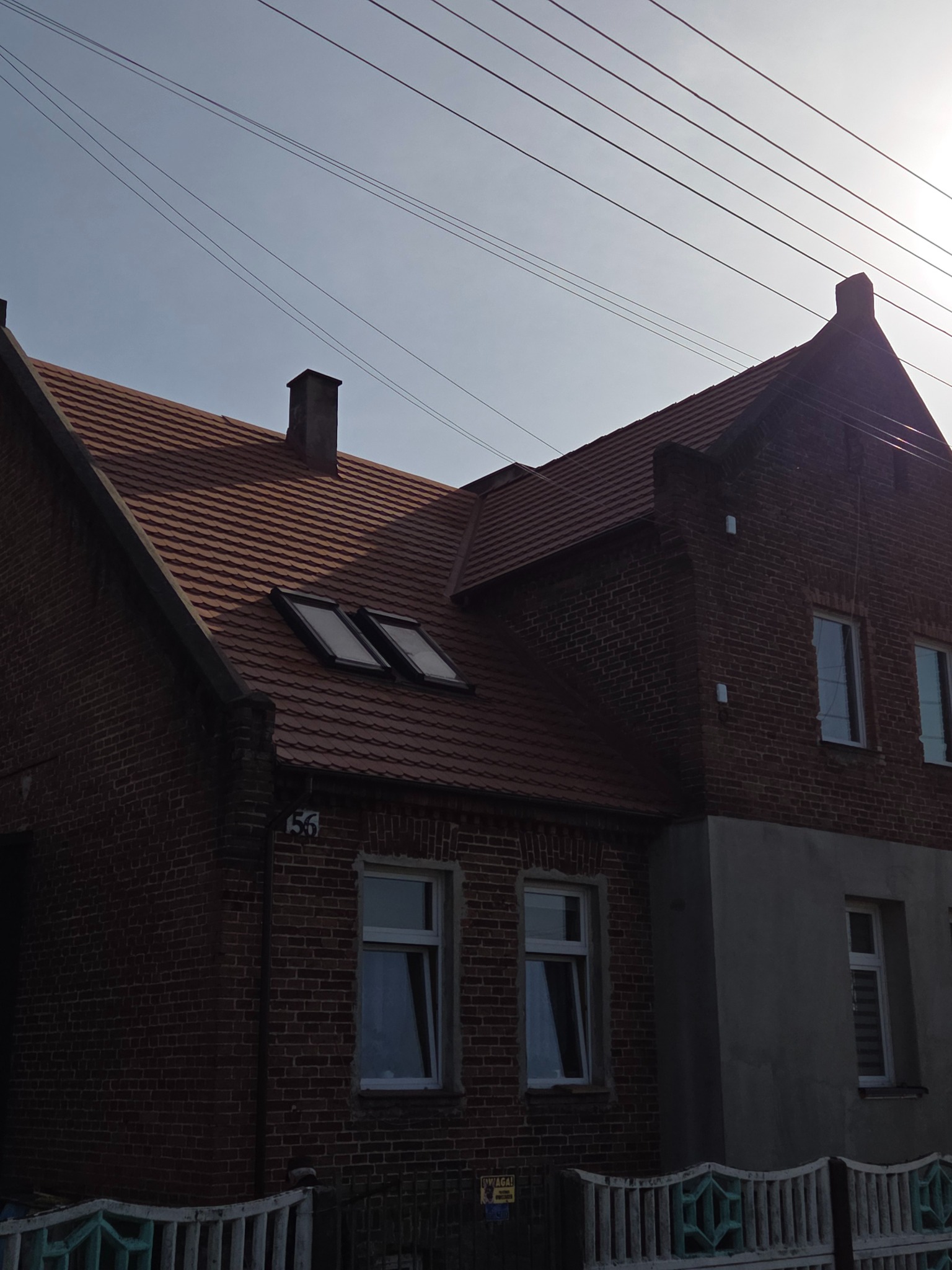 Ceglany dom z nową, czerwoną dachówką i oknami dachowymi. Widoczne przewody elektryczne nad budynkiem. Ujęcie z dołu, w słoneczny dzień.
