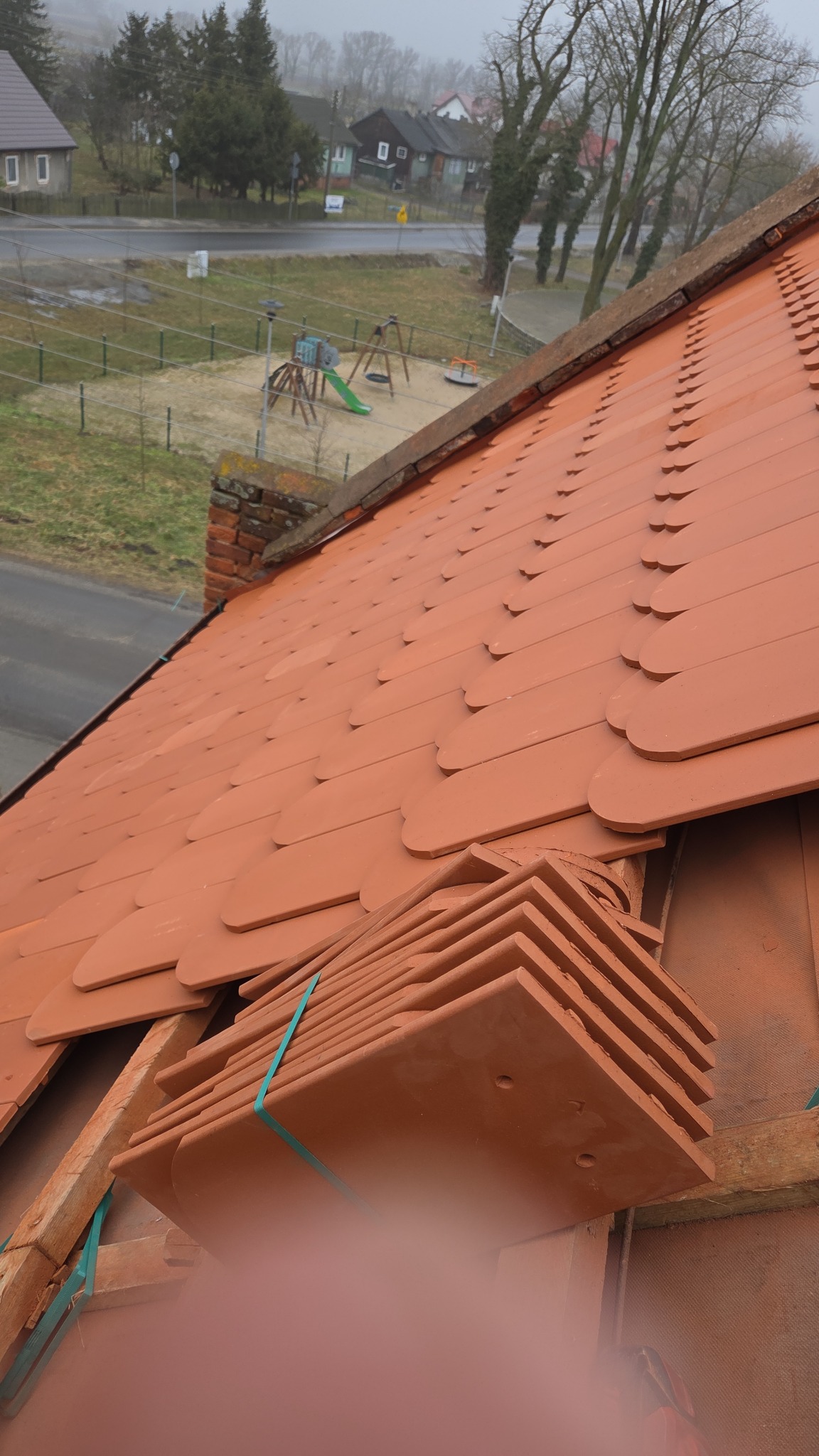 Układanie dachówki ceramicznej na dachu domu jednorodzinnego. Widoczne nowe dachówki w pakietach i fragment dachu pokryty dachówką. W tle plac zabaw.