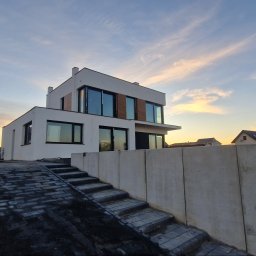 VoltBud - Nowoczesny, dwukondygnacyjny dom z białymi ścianami i dużymi oknami, widziany z dołu, z brukowanym podjazdem i betonowym murem oporowym na pierwszym planie, oświetlony ciepłym światłem zachodu.