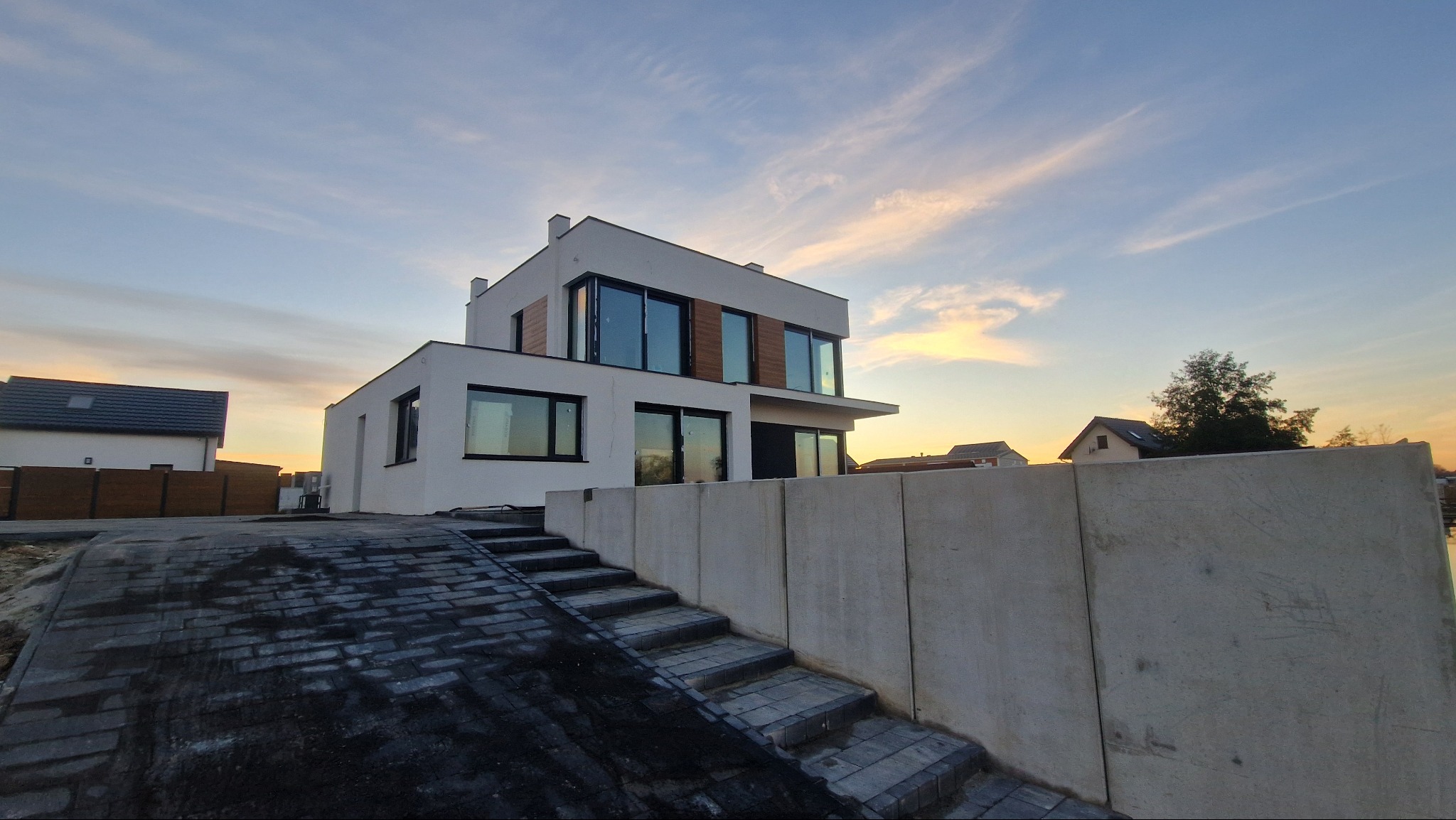 Nowoczesny, dwukondygnacyjny dom z białymi ścianami i dużymi oknami, widziany z dołu, z brukowanym podjazdem i betonowym murem oporowym na pierwszym planie, oświetlony ciepłym światłem zachodu.