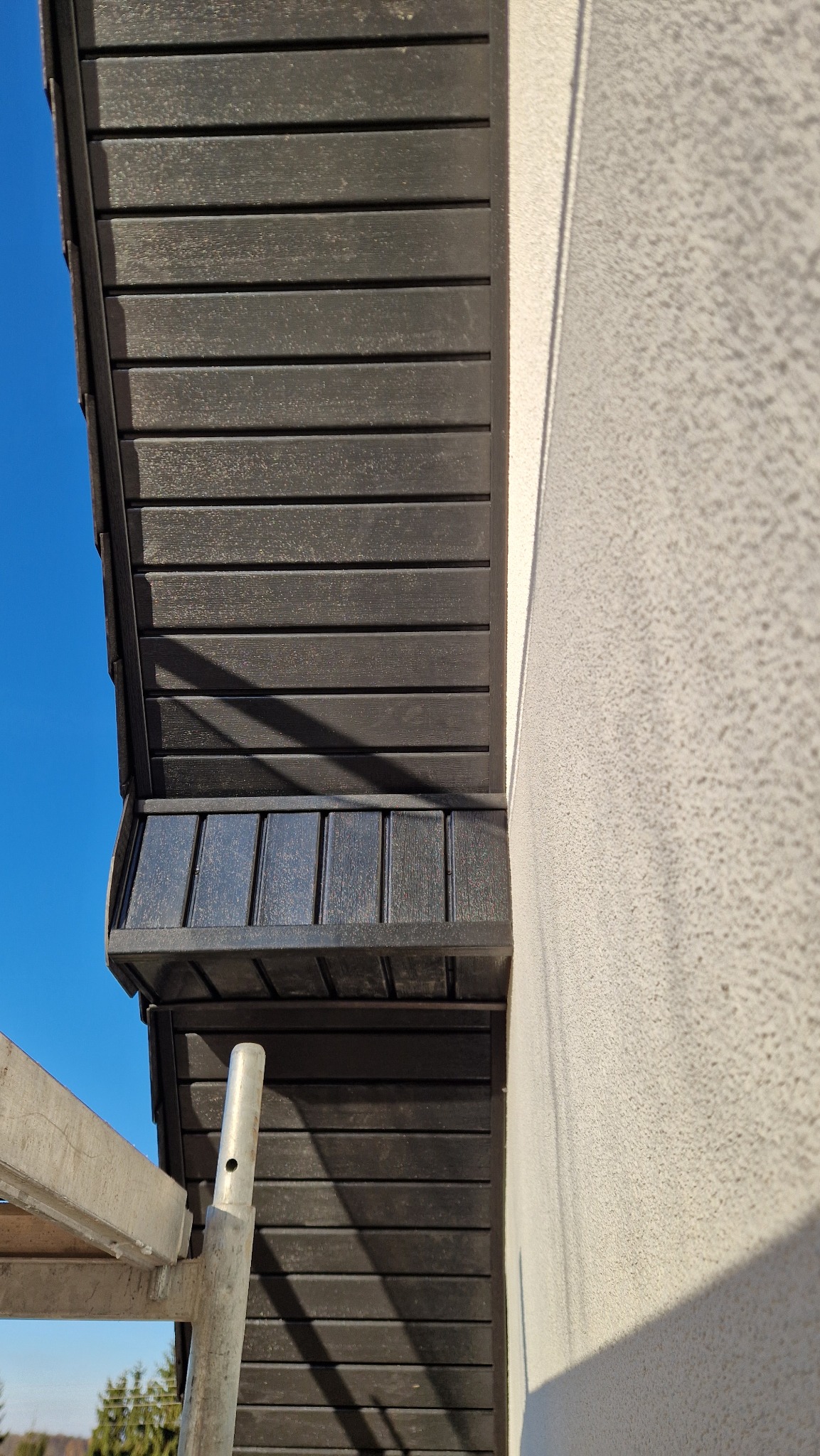 Ciemna podbitka dachowa z desek kompozytowych, widok z dołu, fragment rusztowania i elewacja budynku w tle, błękitne niebo. Ujęcie z dołu.