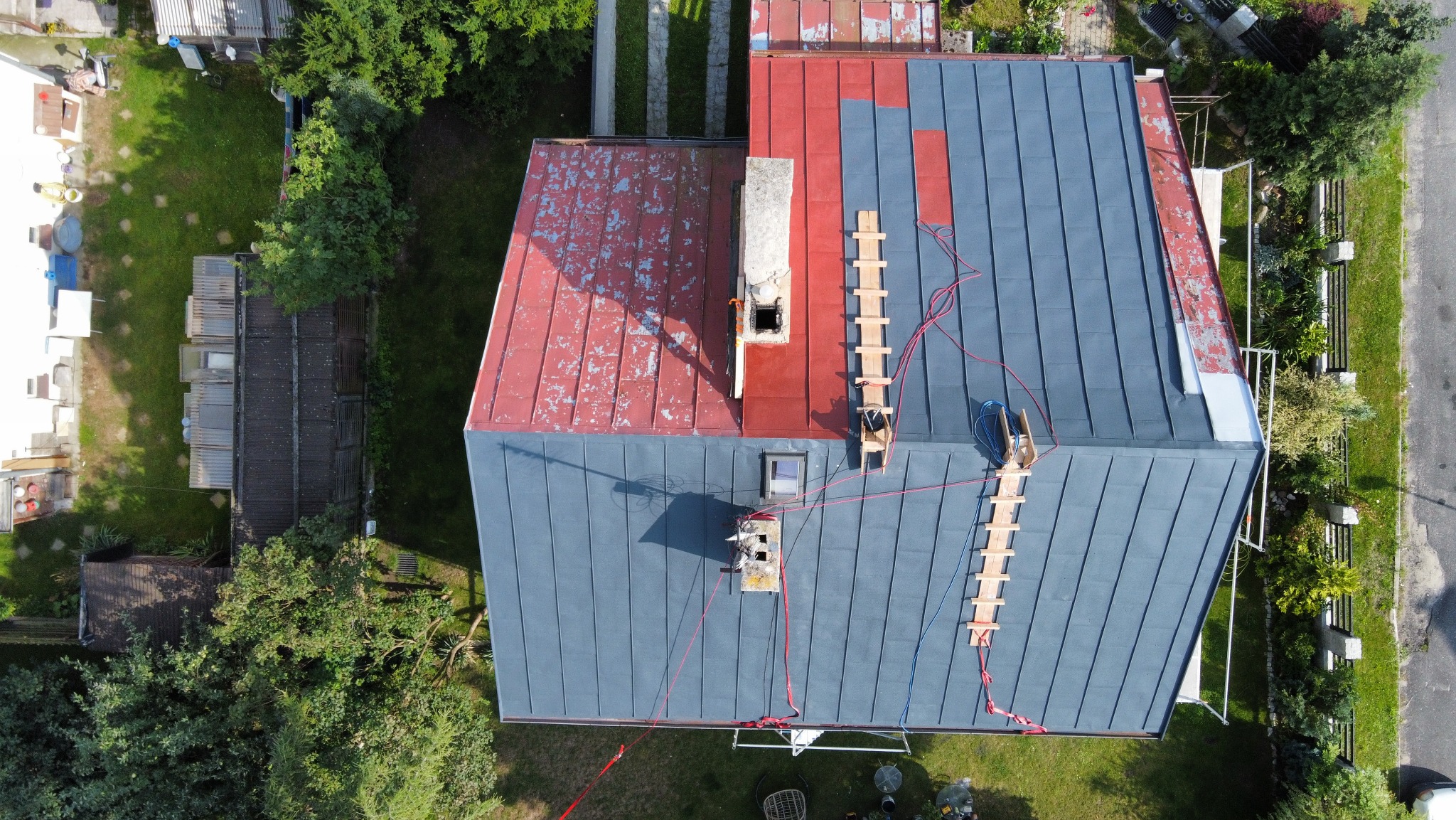Zdjęcie z drona przedstawia remontowany dach domu: część dachu pomalowana na szaro, część w kolorze czerwonym. Widoczne rusztowania i narzędzia. Otoczenie to zieleń.