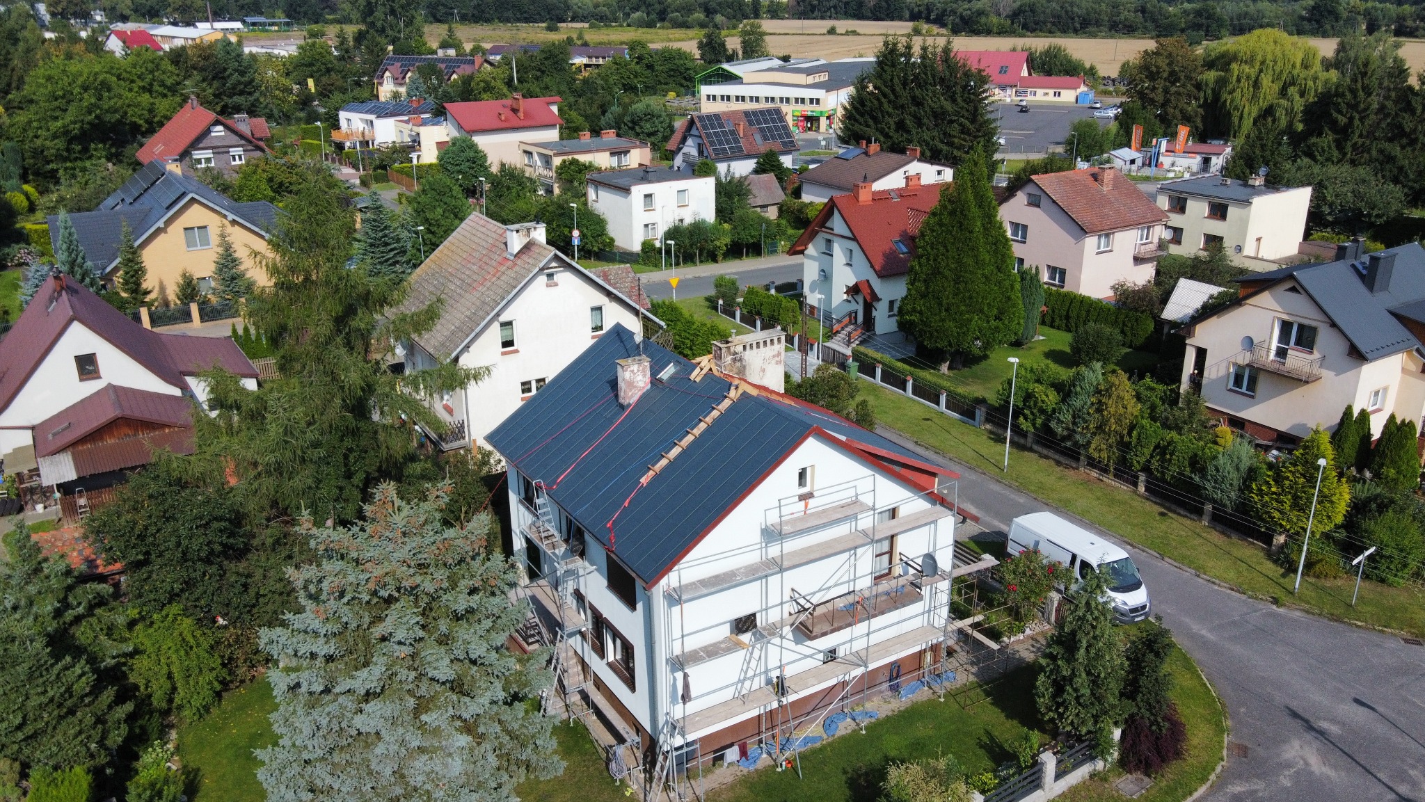 Dom z nowym, ciemnym dachem w trakcie montażu, otoczony rusztowaniami. Widok z góry na budynek w zabudowie jednorodzinnej, z zielonym otoczeniem i zaparkowanym busem.