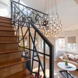 Wnętrze domu z drewnianymi schodami, czarną, geometryczną balustradą i nowoczesną lampą kulistą, widok z dołu na salon z beżową sofą i oknami.