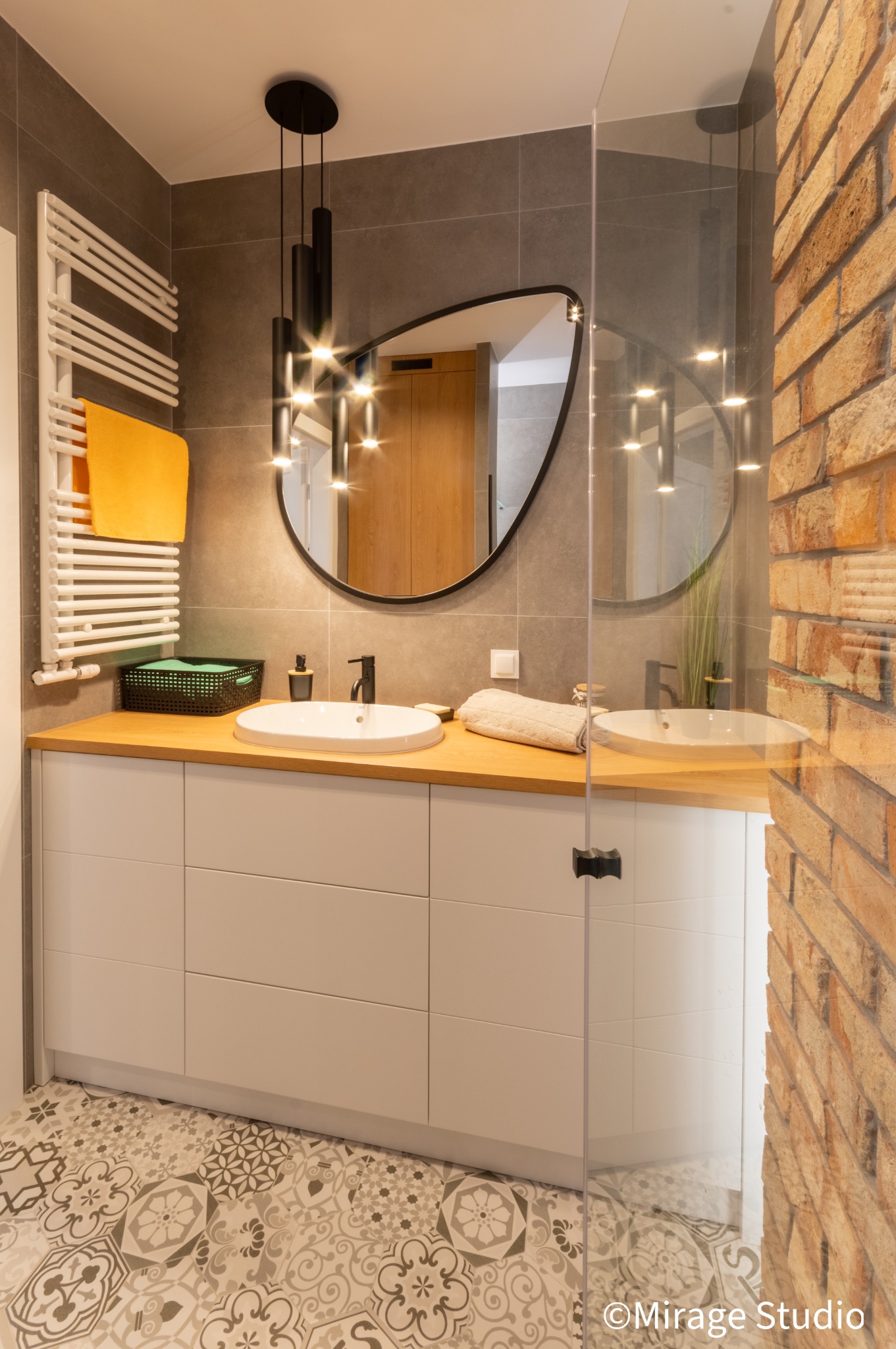 Nowoczesna łazienka z podwójną umywalką na drewnianym blacie, białe szafki, dekoracyjna podłoga, ceglana ściana za szklaną kabiną prysznicową i wisząca lampa z czarnymi cylindrami.