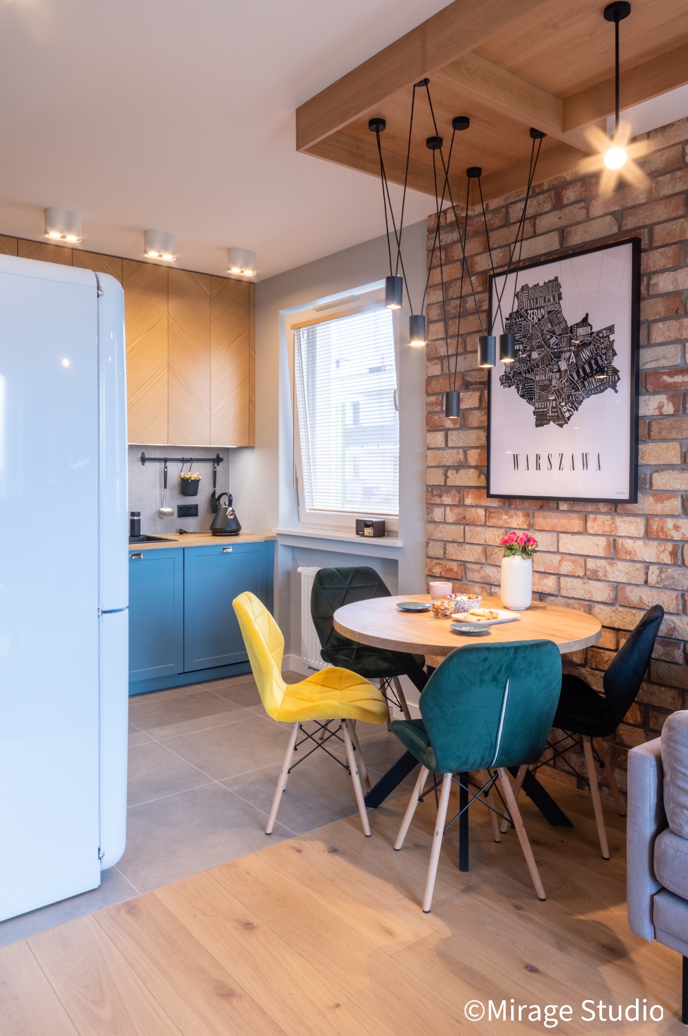 Jasne wnętrze kuchni z jadalnią: niebieskie szafki, drewniane blaty, okrągły stół z kolorowymi krzesłami, ceglana ściana z plakatem mapy Warszawy, wiszące lampy.