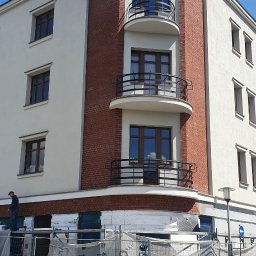 LIS BARTŁOMIEJ ZAKŁAD REMONTOWO-BUDOWLANY - Budynek z elewacją w trakcie prac. Widoczne rusztowania, ocieplenie styropianem i pracownik na drabinie. Część budynku wykończona czerwoną cegłą.