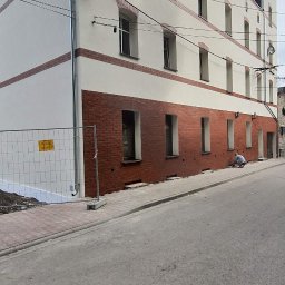 LIS BARTŁOMIEJ ZAKŁAD REMONTOWO-BUDOWLANY - Odnowiona elewacja budynku z ceglanym pasem u dołu i białymi ścianami powyżej. Widoczny pracownik przy chodniku, metalowe barierki i fragment ulicy.