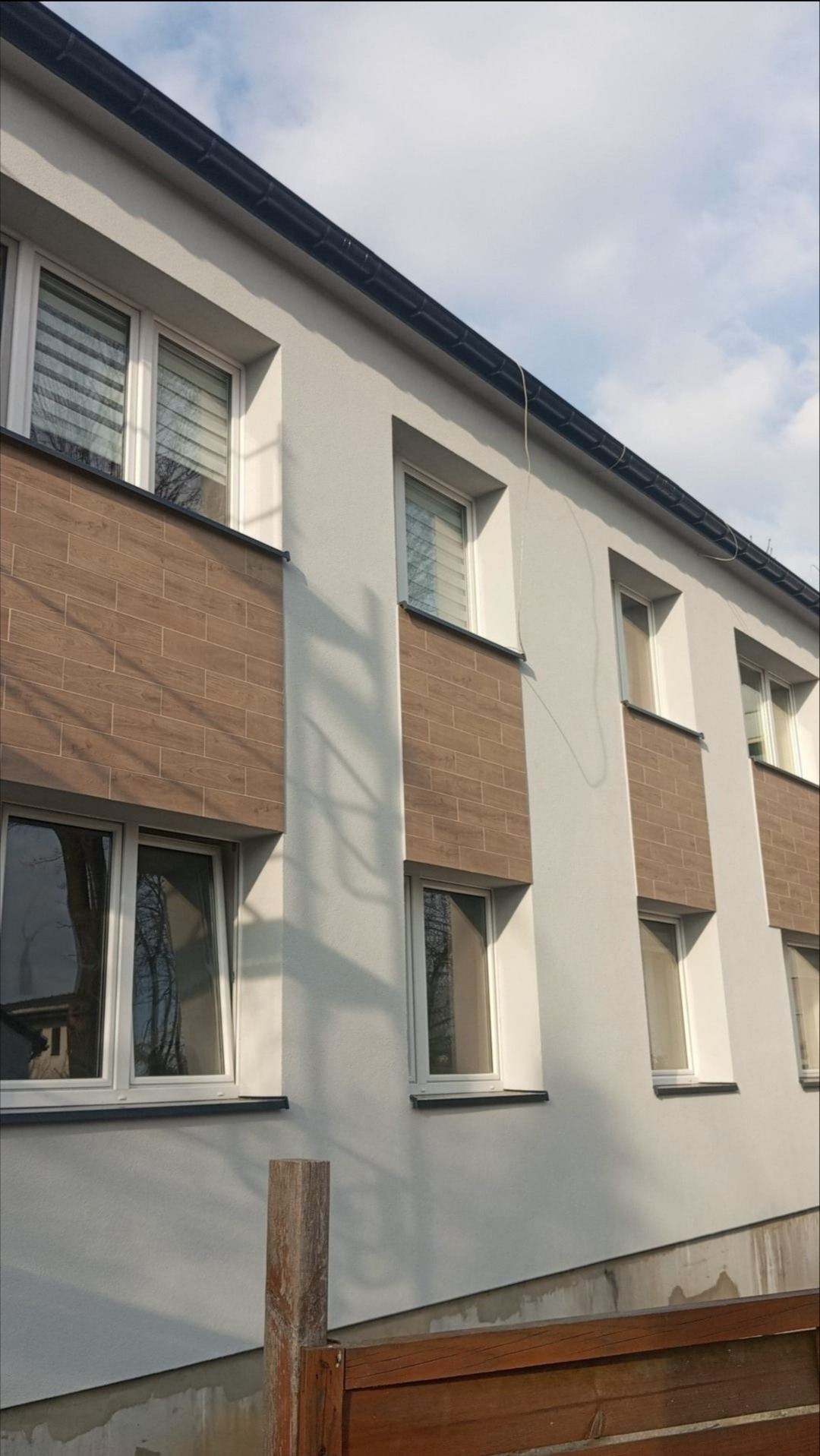 Elewacja budynku z oknami i dekoracyjnymi panelami imitującymi drewno, widziana pod kątem. Widoczne fragmenty ogrodzenia i niebieskie niebo.