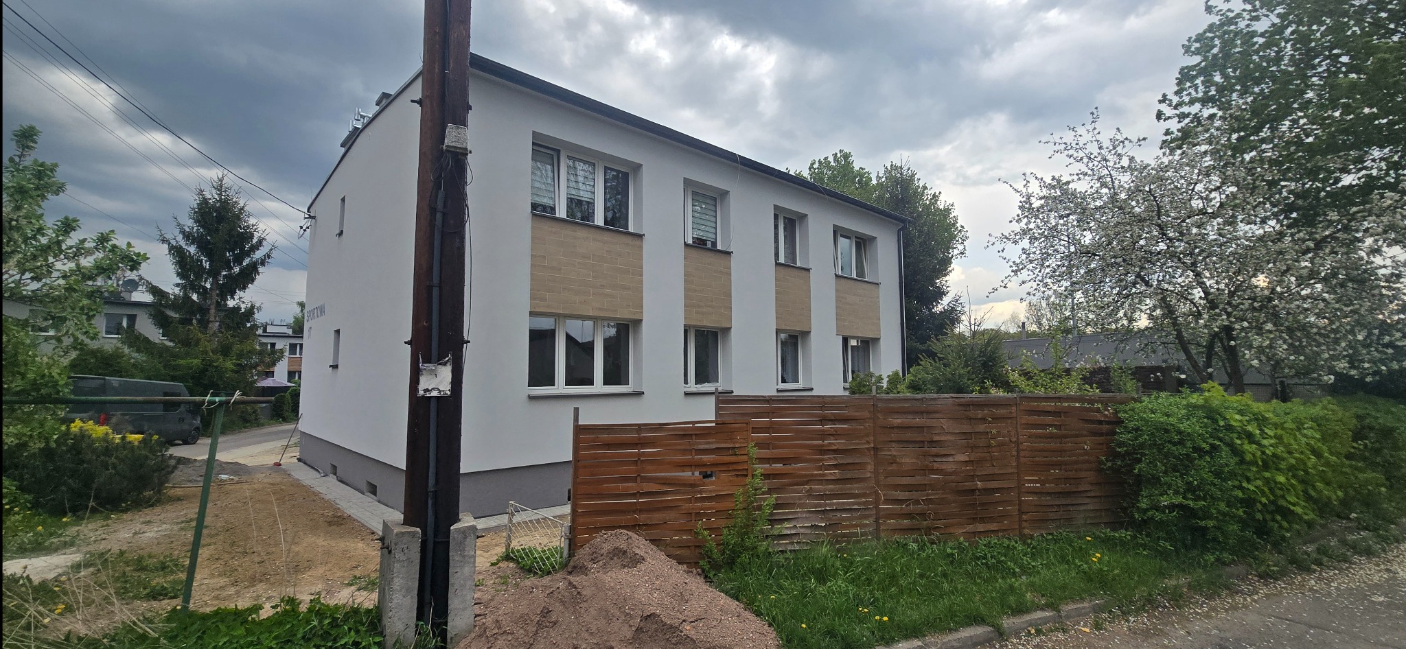 Dwupiętrowy budynek po renowacji elewacji, z białymi ścianami i drewnianymi panelami. Widoczny nowy płot i przygotowany teren pod kostkę. Pochmurne niebo w tle.