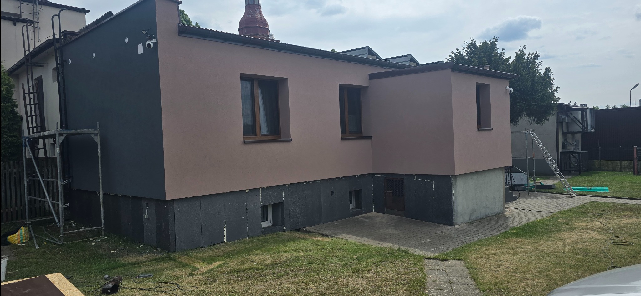 Dwukolorowa elewacja domu jednorodzinnego w trakcie prac. Część budynku ocieplona styropianem, część pomalowana na szaro. Widoczne rusztowania i drabina.