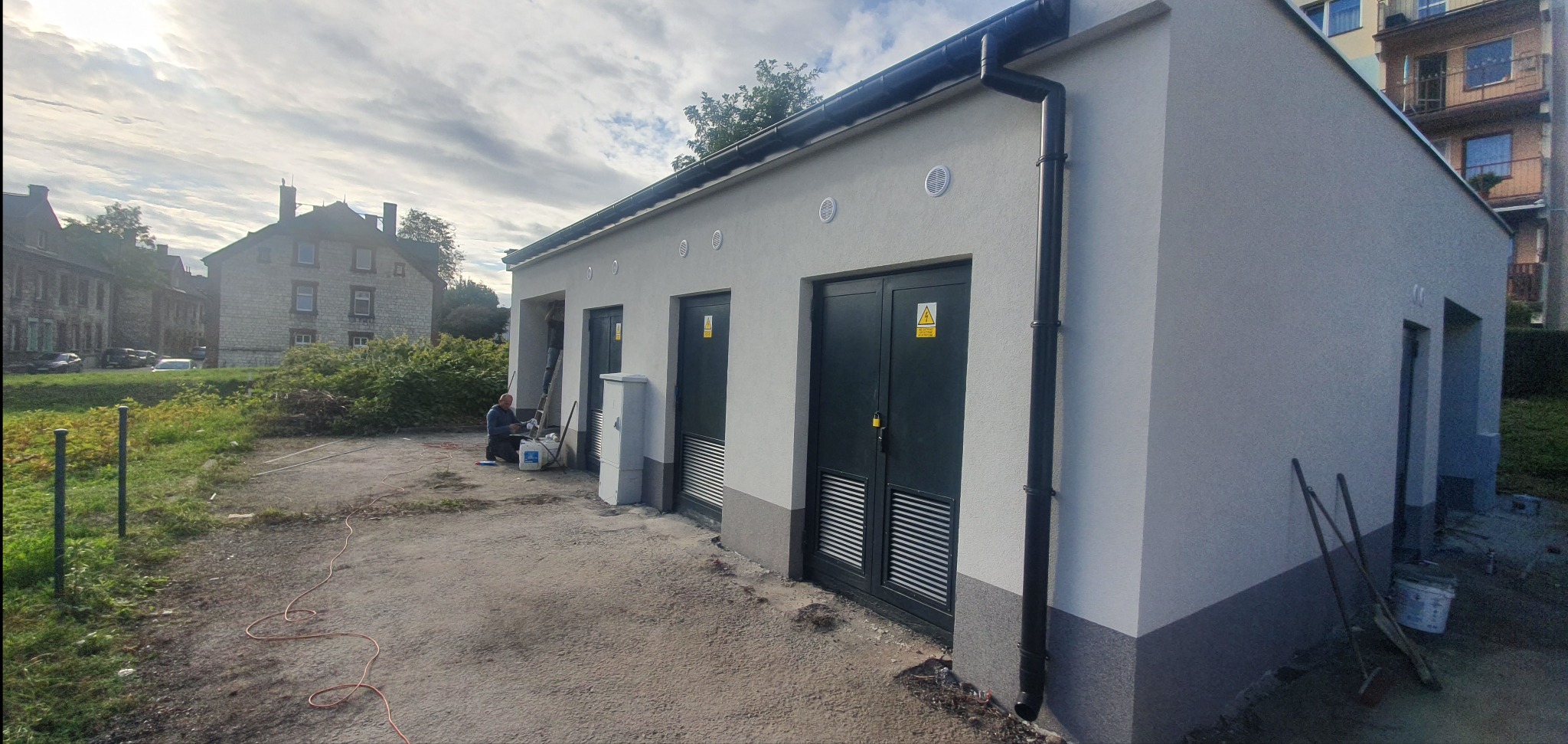Szara elewacja budynku transformatorowego z czarnymi drzwiami i wentylacją. Pracownik maluje wnękę. Widoczne narzędzia i wiadro z farbą. Budynek otoczony zielenią.