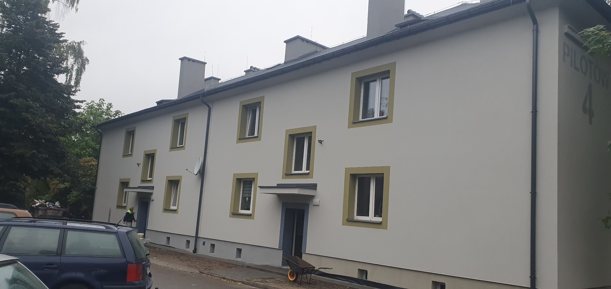 Odnowiona elewacja dwupiętrowego budynku z szarymi oknami i numerem 'Pilotów 4' na rogu. Widoczny fragment samochodu i pracownik przy wejściu.