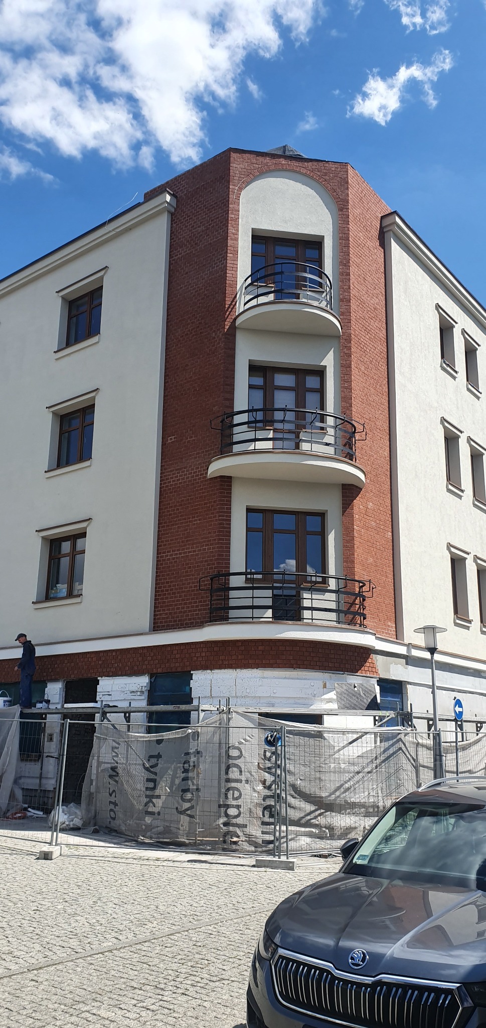 Budynek z elewacją w trakcie prac. Widoczne rusztowania, ocieplenie styropianem i pracownik na drabinie. Część budynku wykończona czerwoną cegłą.