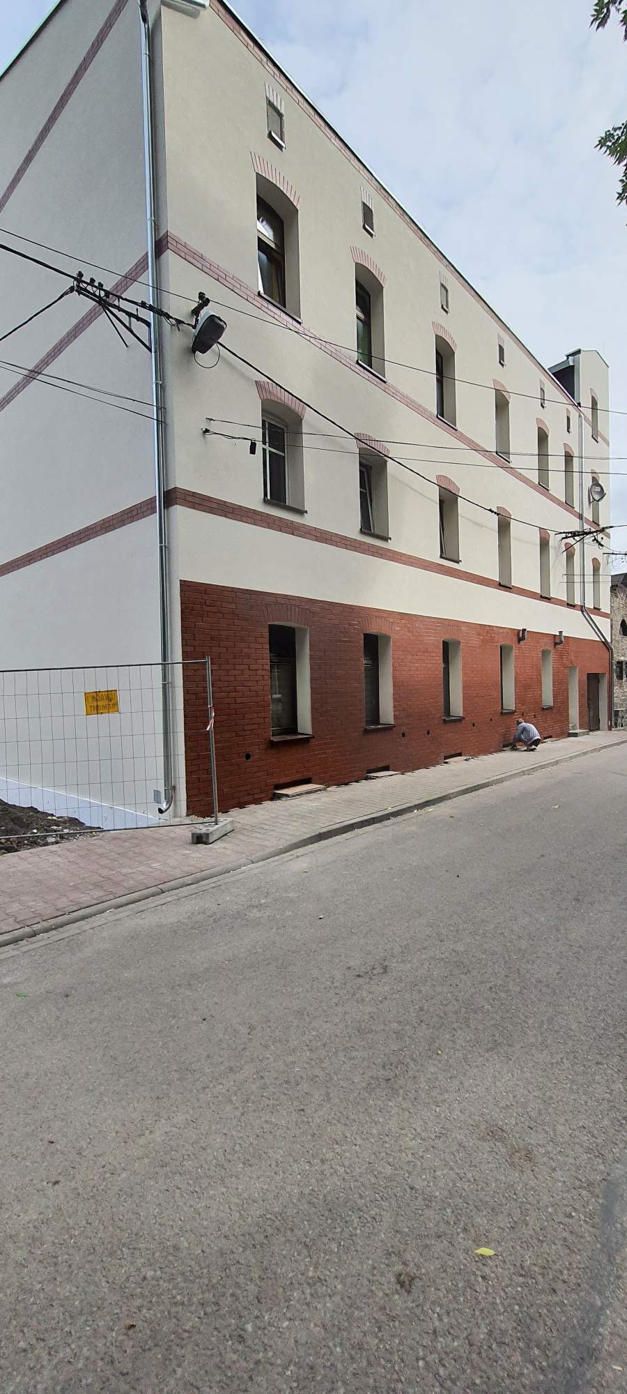 Odnowiona elewacja budynku z ceglanym pasem u dołu i białymi ścianami powyżej. Widoczny pracownik przy chodniku, metalowe barierki i fragment ulicy.
