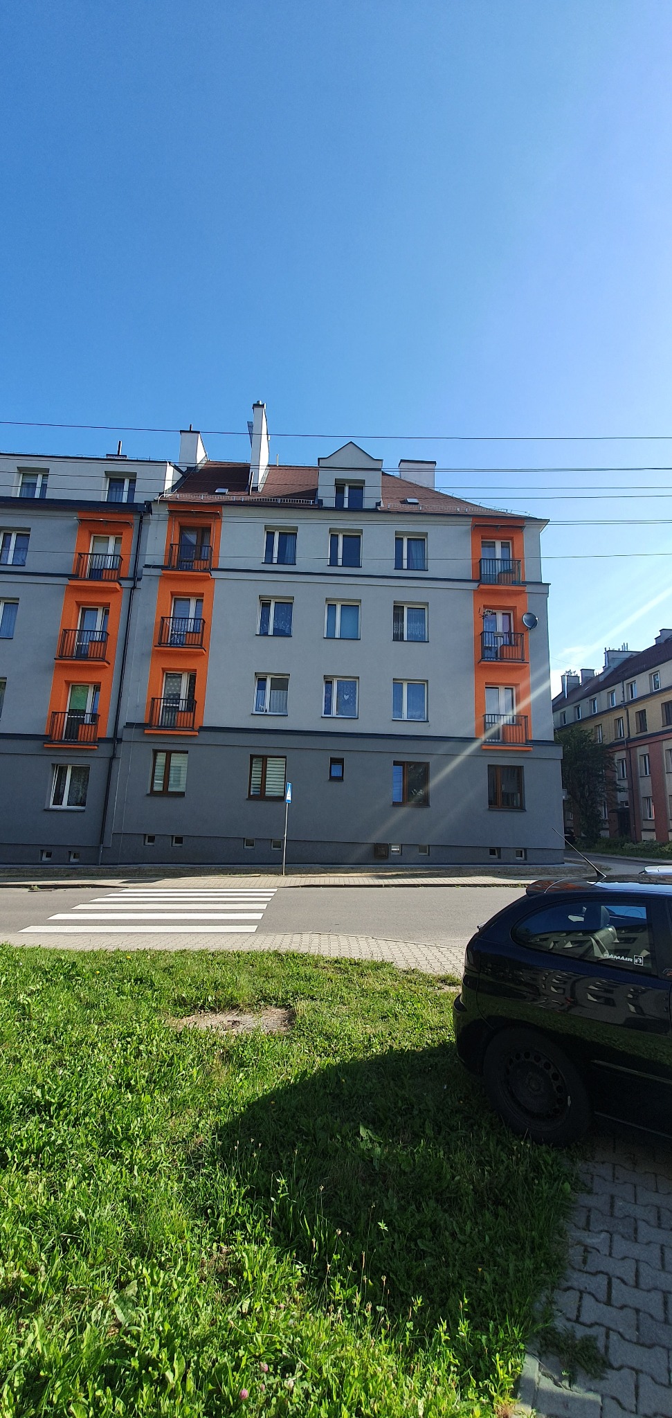 Wielopiętrowy budynek po renowacji z pomarańczowymi akcentami w Dąbrowie Górniczej, widoczny z poziomu ulicy. Na pierwszym planie trawnik i fragment samochodu.
