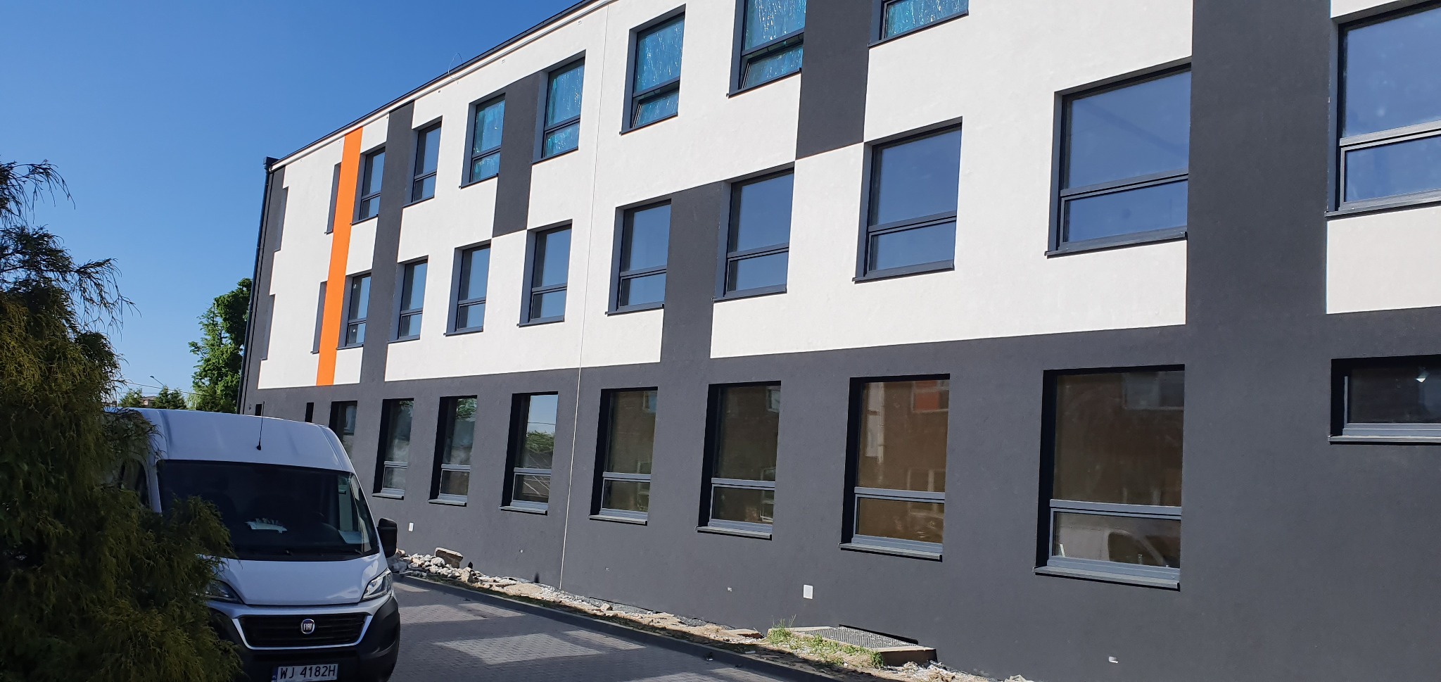 Nowoczesna elewacja budynku z akcentem pomarańczowym, biało-szara kolorystyka, widoczny fragment chodnika i samochód dostawczy. Słoneczny dzień.