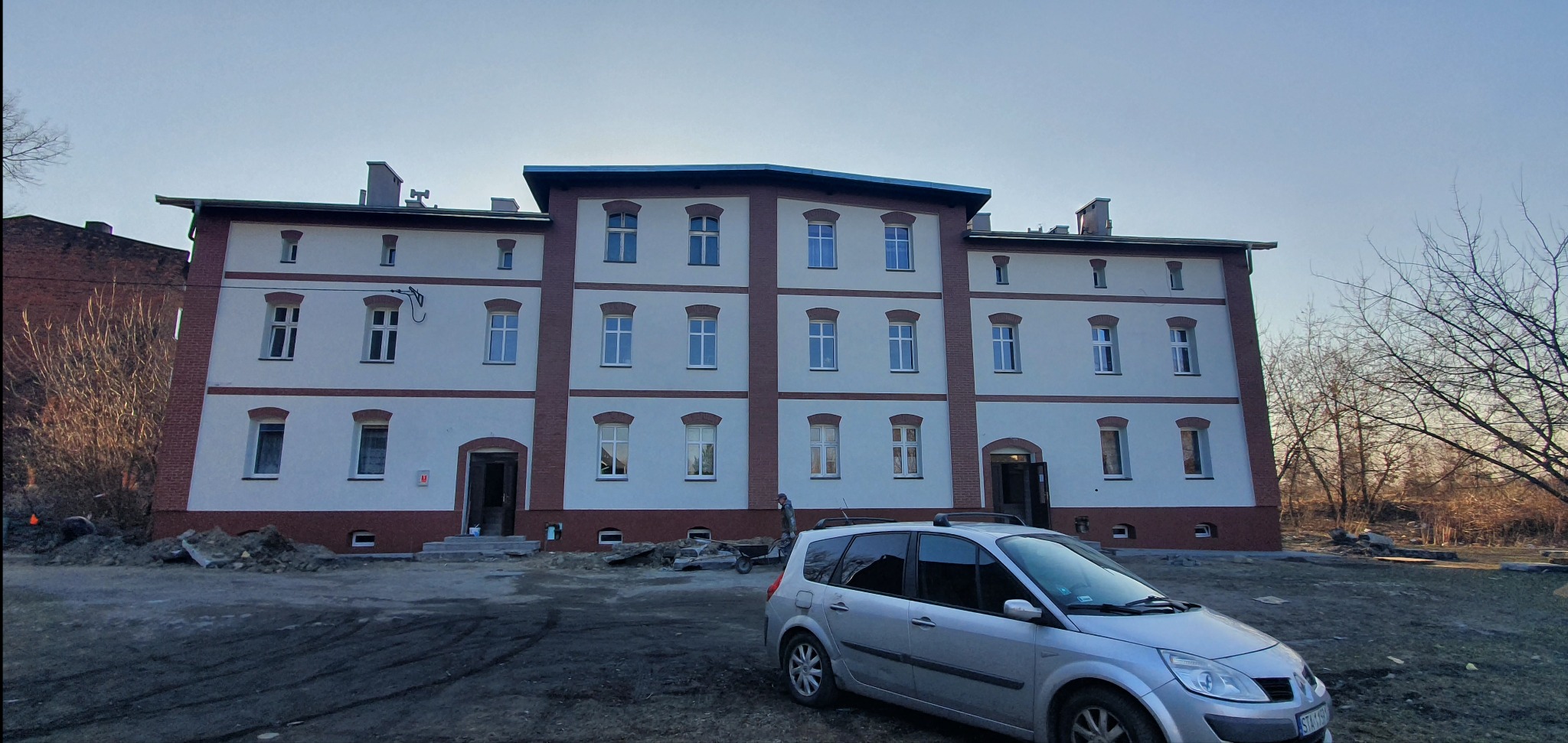 Odnowiona elewacja budynku z białymi ścianami i ceglanymi akcentami. Widoczny samochód i teren po pracach budowlanych. Ujęcie w pochmurny dzień.