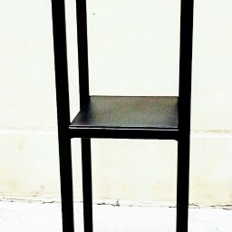 Fenix Michał Tupta, Rafał łapa Spòłka cywilna - Czarny, metalowy regał z dwiema półkami, spawany z profili kwadratowych, ustawiony na jasnym tle ściany i podłogi.