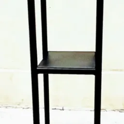 Czarny, metalowy regał z dwiema półkami, spawany z profili kwadratowych, ustawiony na jasnym tle ściany i podłogi.