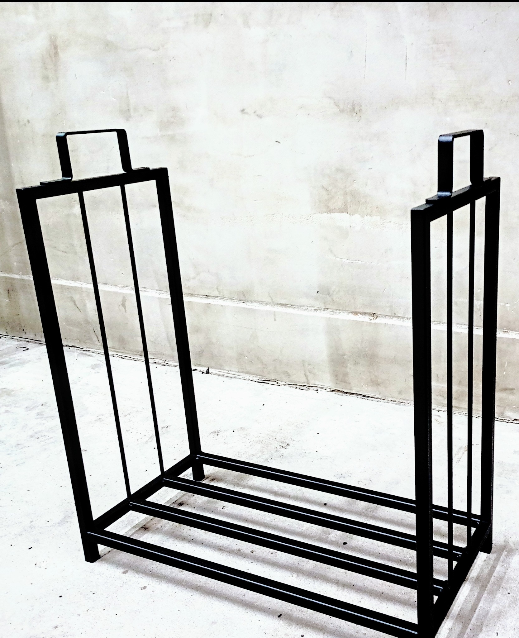 Czarny, metalowy stojak na drewno kominkowe z uchwytami, spawana konstrukcja na tle szarej, betonowej ściany.