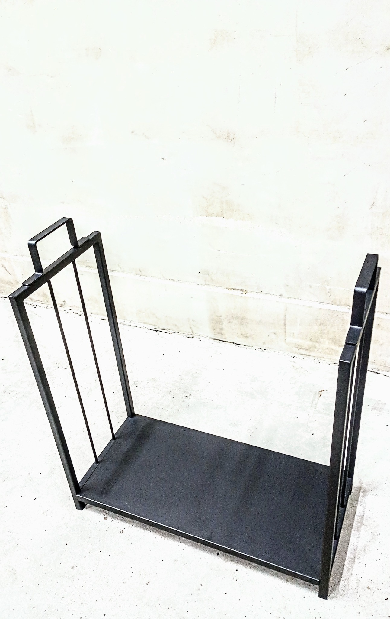 Czarny, minimalistyczny stojak na drewno kominkowe wykonany z metalowych profili o prostokątnym przekroju, ustawiony na betonowej posadzce i sfotografowany na tle jasnej ściany.