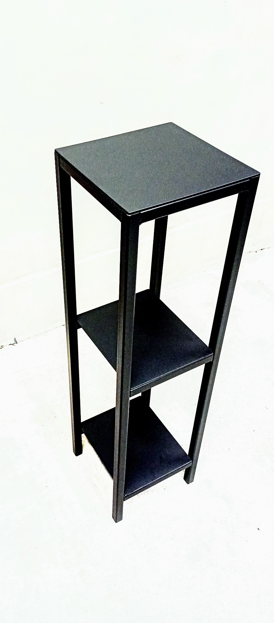 Czarny, metalowy, trzypoziomowy stolik kawowy o minimalistycznym designie, stojący na białym tle.