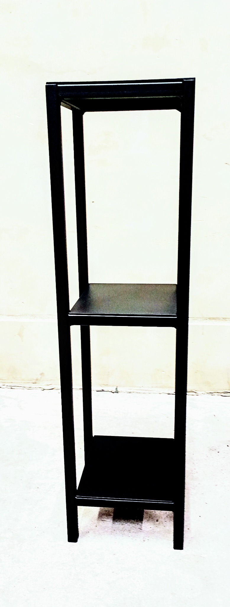 Czarny, metalowy regał z dwiema półkami, spawany z profili kwadratowych, ustawiony na jasnym tle ściany i podłogi.