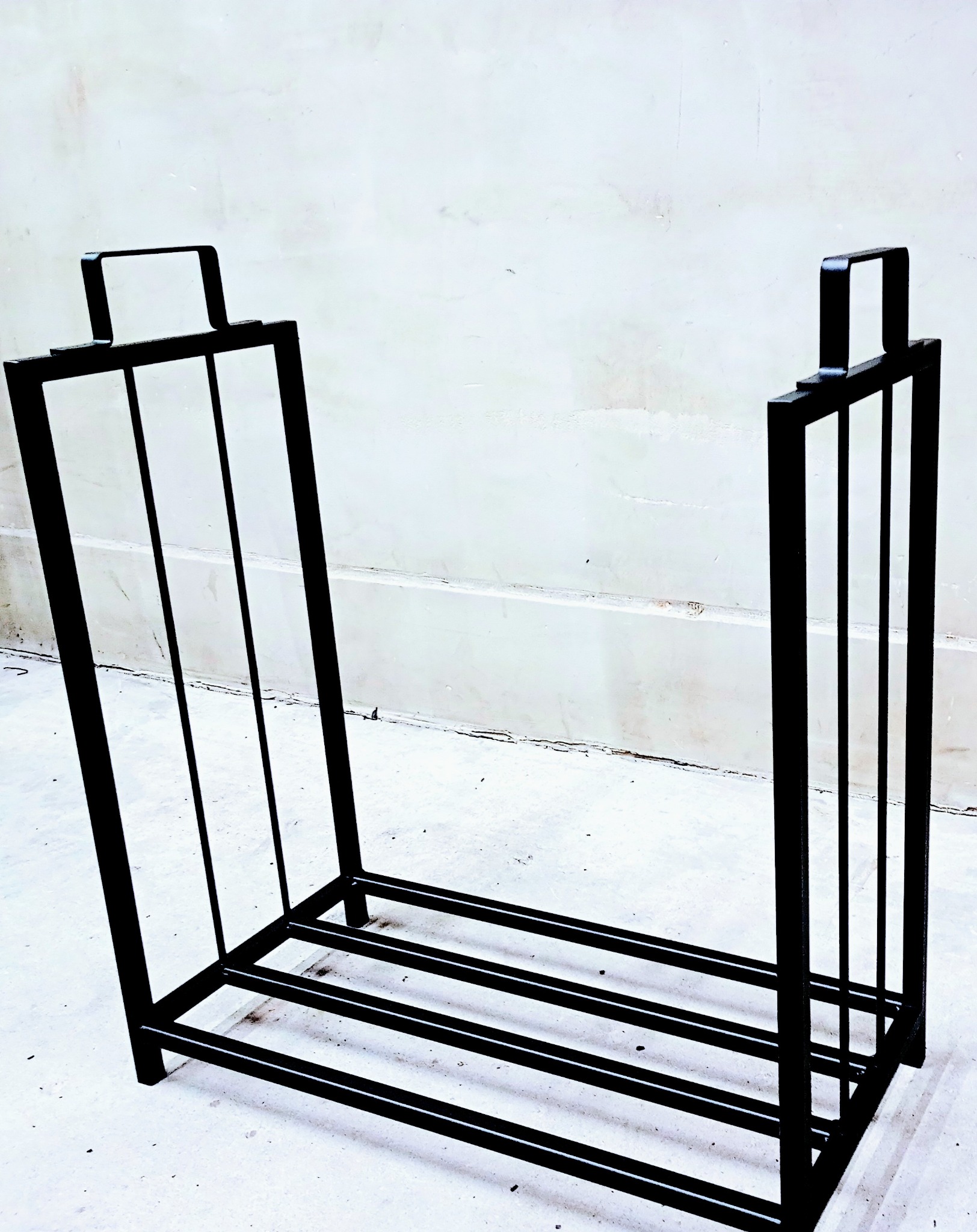 Metalowy stojak na drewno kominkowe z czarnych, prostych profili, widoczny na tle jasnej ściany.