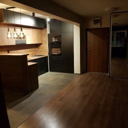 Mountain - Nowoczesna kuchnia z drewnianymi frontami i czarnymi szafkami, wykończona złotą armaturą i lampami wiszącymi, widok z korytarza z drewnianą podłogą i lustrem.