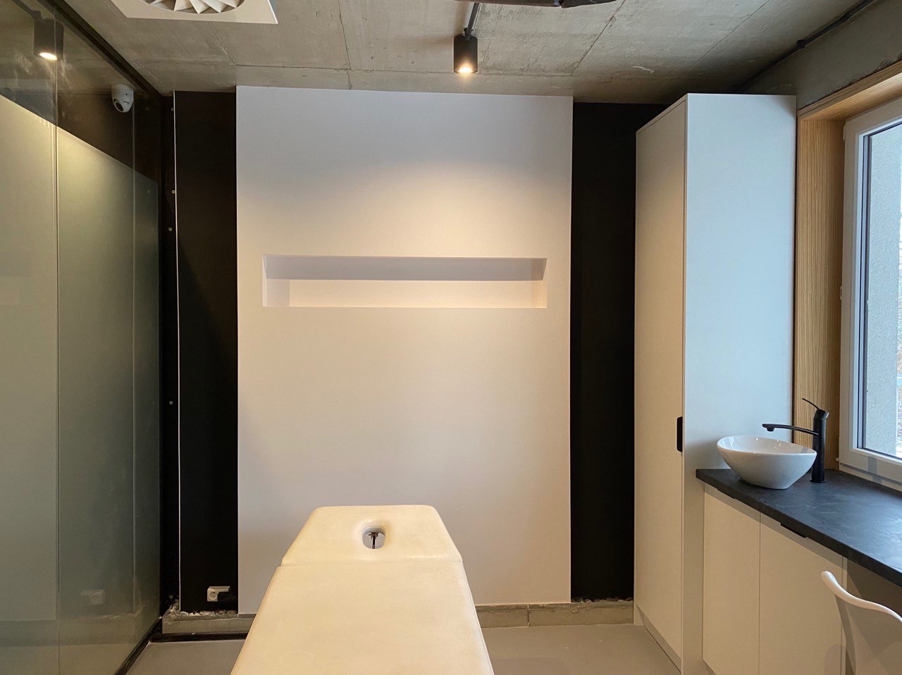 Nowoczesna łazienka z białymi ścianami, wnęką z oświetleniem, umywalką nablatową na czarnym blacie i betonowym sufitem z oświetleniem punktowym.