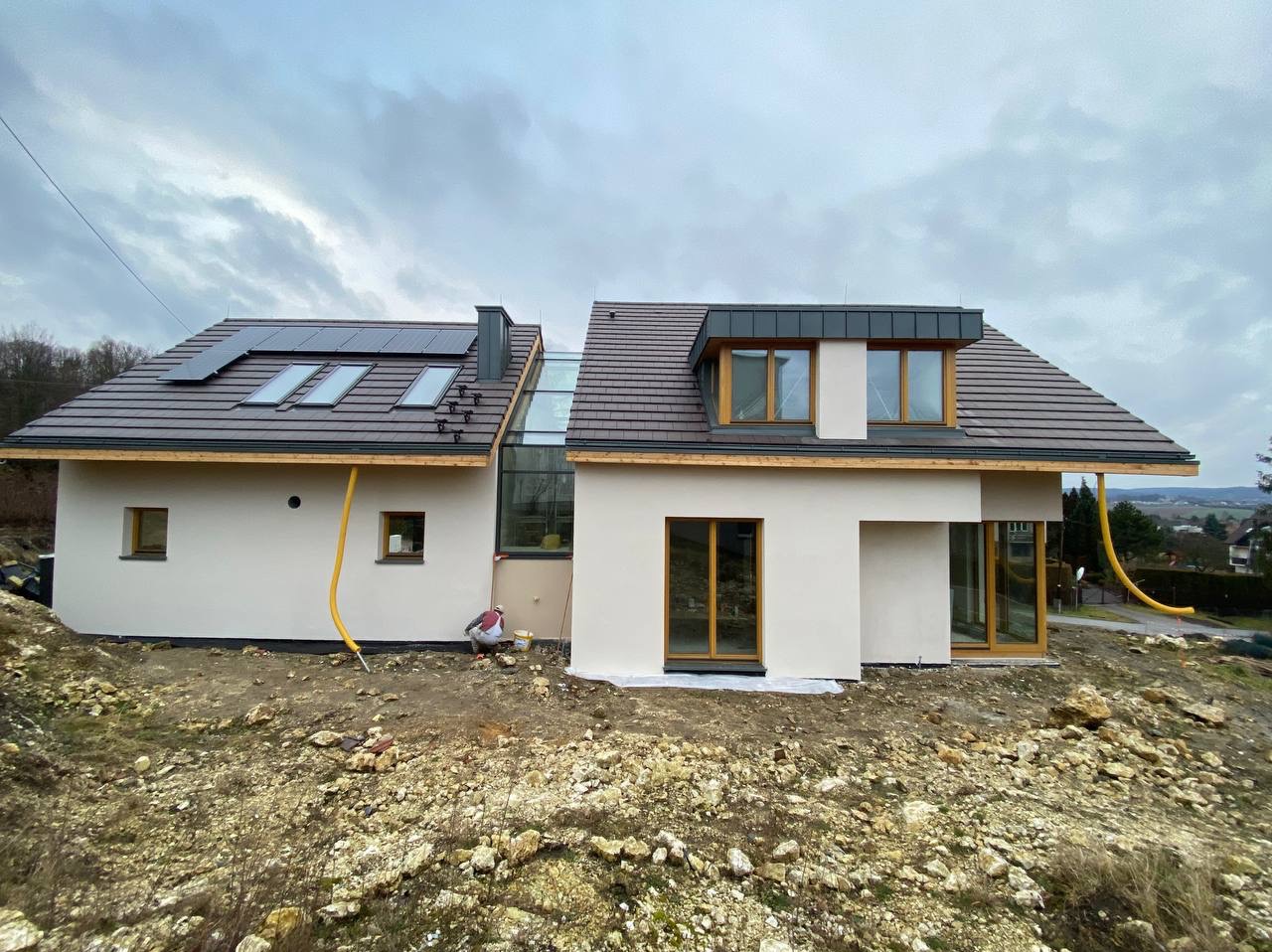 Dwukondygnacyjny dom jednorodzinny z panelami słonecznymi i oknami dachowymi, świeżo otynkowany na biało, z żółtym wężem odprowadzającym wodę i pracownikiem w kasku przy ścianie, w trakcie prac...