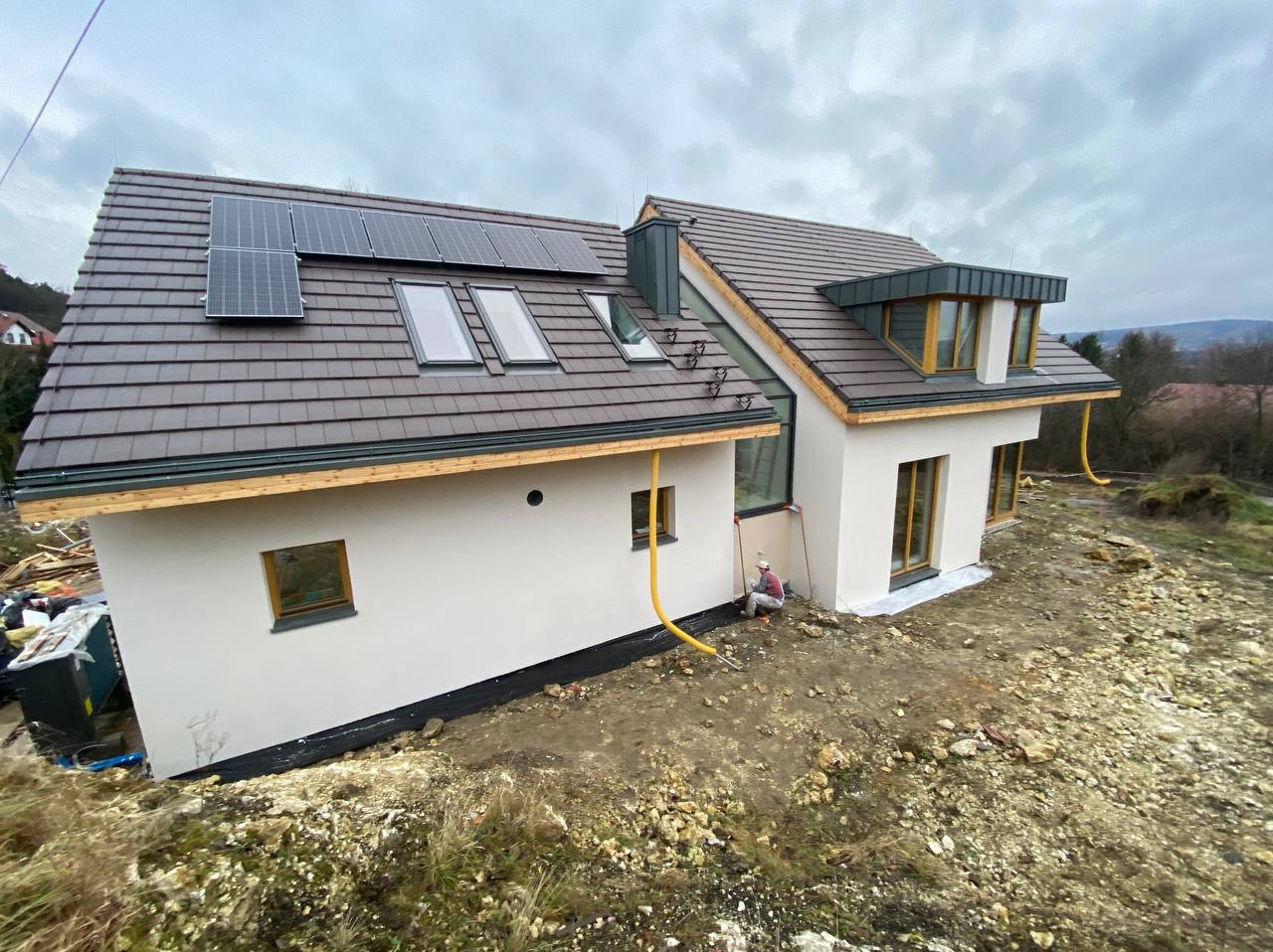 Nowoczesny dom jednorodzinny z brązowym dachem, panelami słonecznymi i oknami dachowymi, elewacja w trakcie wykończenia, widoczny pracownik w kombinezonie przy ścianie, żółta rura odprowadzająca...