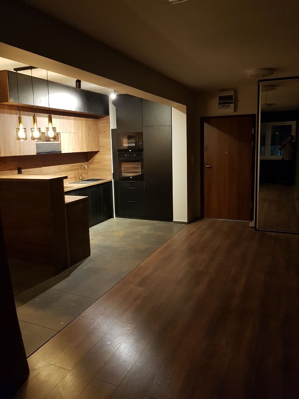 Nowoczesna kuchnia z drewnianymi frontami i czarnymi szafkami, wykończona złotą armaturą i lampami wiszącymi, widok z korytarza z drewnianą podłogą i lustrem.