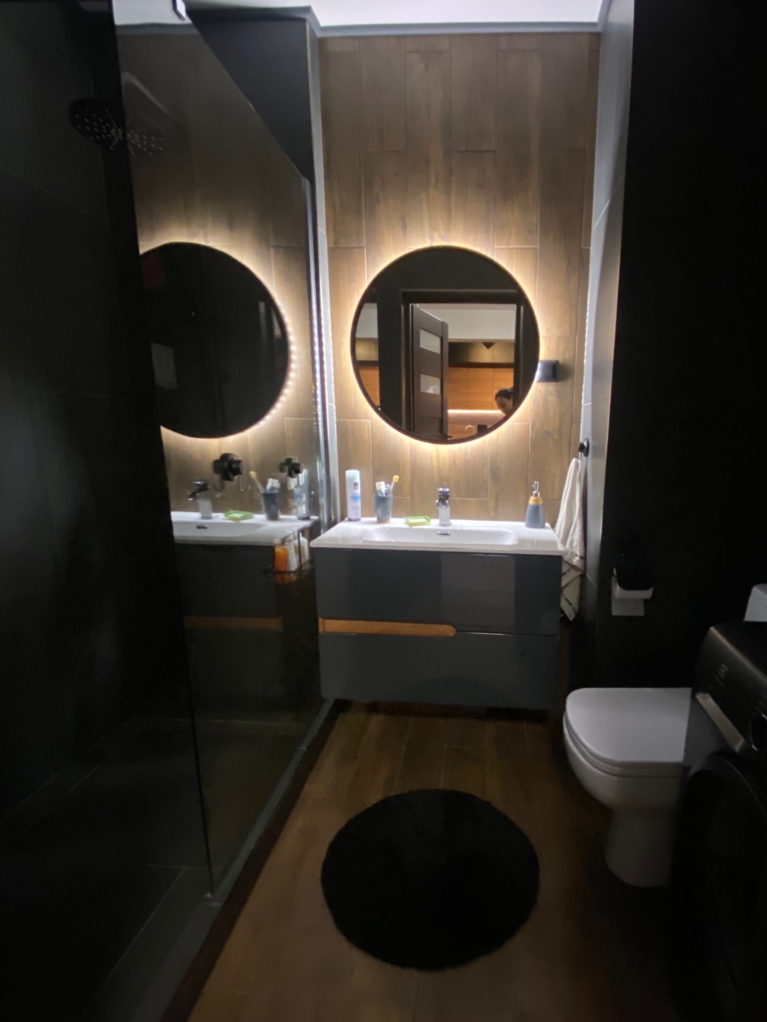 Nowoczesna łazienka w Krakowie z podświetlanym okrągłym lustrem nad szarą szafką umywalkową, widoczna kabina prysznicowa z czarnymi płytkami oraz drewnopodobne płytki na ścianie.