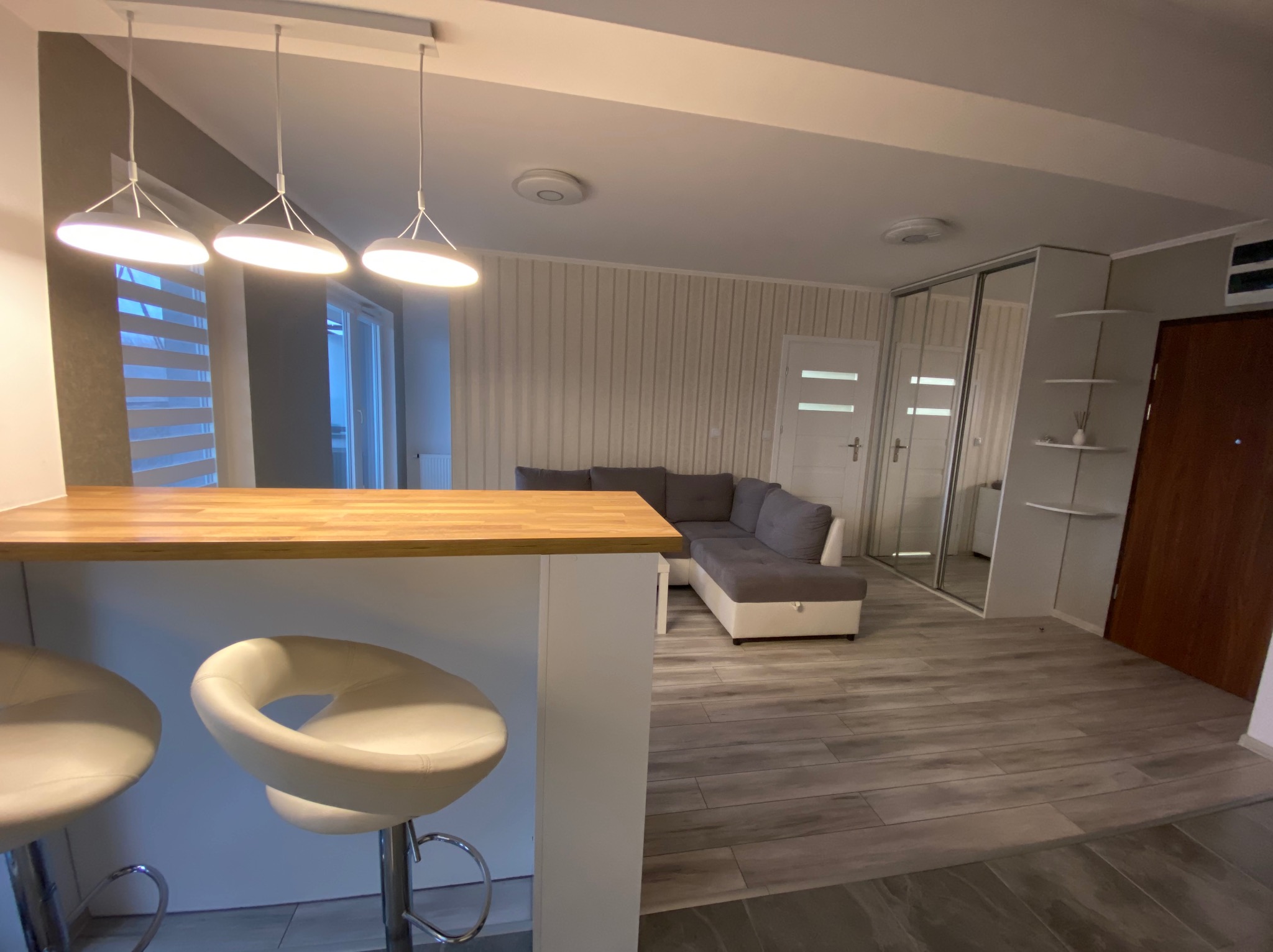 Nowoczesne wnętrze: jasna kuchnia z drewnianym blatem, białe hokery, szara sofa narożna i podłoga z płytek imitujących drewno.