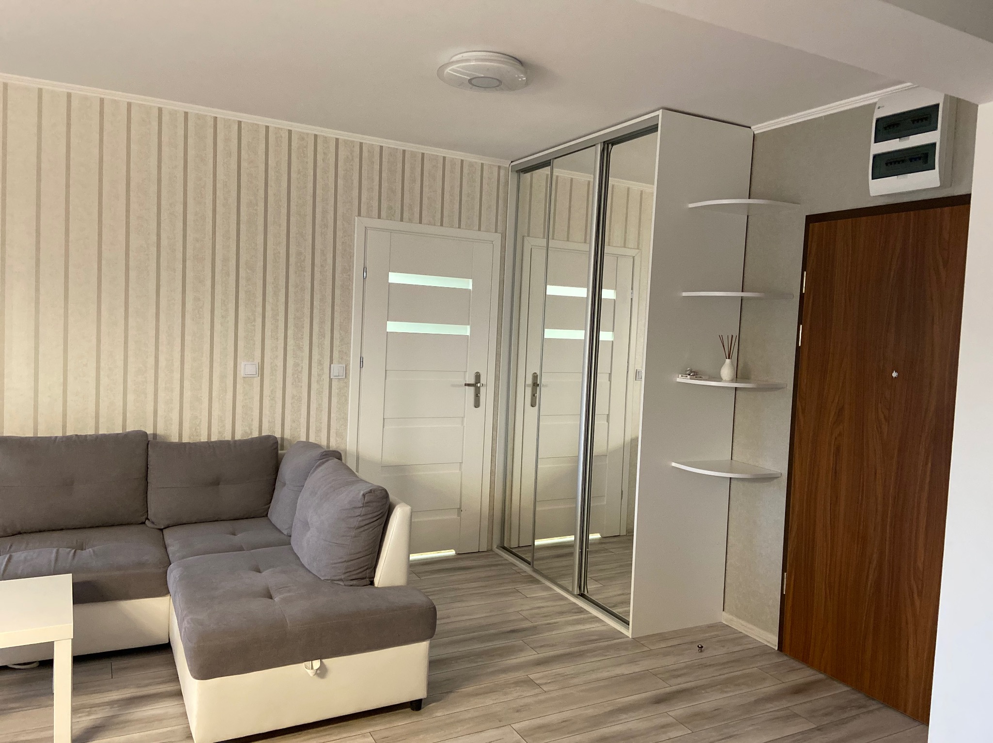Wnętrze pokoju po remoncie: Szary narożnik, biały stolik kawowy, szafa z lustrzanymi drzwiami przesuwnymi, białe drzwi z przeszkleniami, ściany w beżowe pionowe pasy, drewniane panele podłogowe.