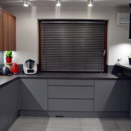 IdealForm Natalia Gosztyła - Nowoczesna kuchnia w szarościach z drewnianymi akcentami, minimalistyczne oświetlenie punktowe i wiszące lampy, okno z żaluzjami, AGD na blacie.
