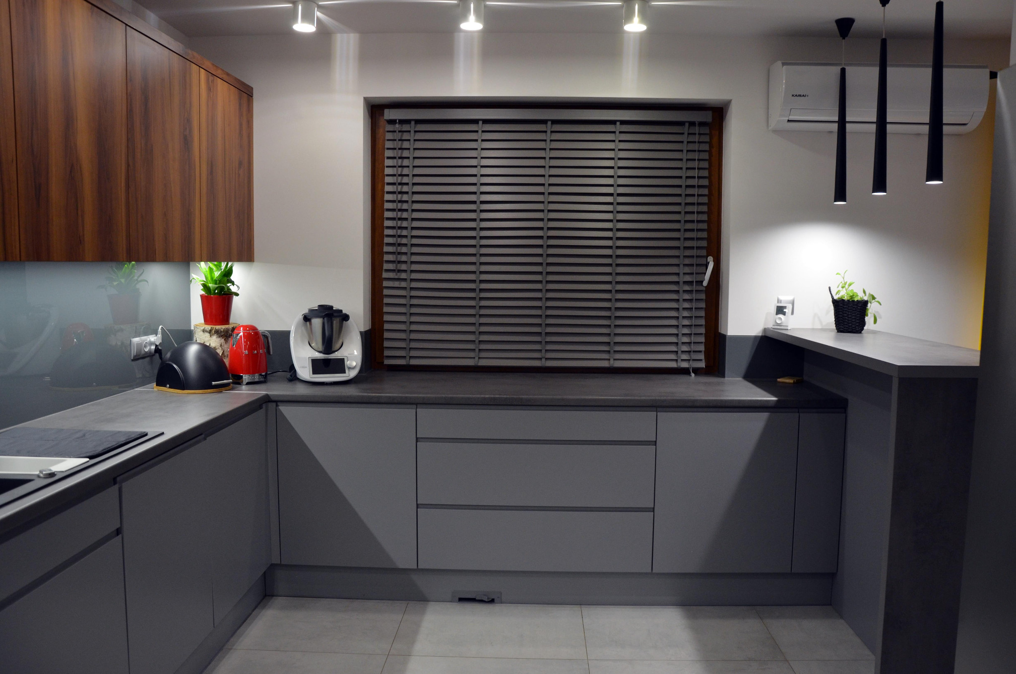 Nowoczesna kuchnia w szarościach z drewnianymi akcentami, minimalistyczne oświetlenie punktowe i wiszące lampy, okno z żaluzjami, AGD na blacie.
