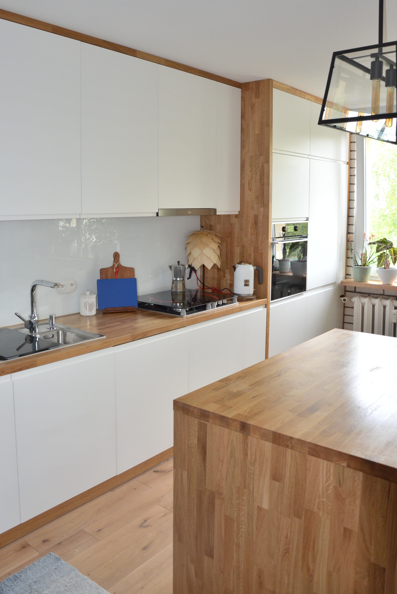 Jasna kuchnia z białymi szafkami i drewnianym blatem, na którym stoi moka ekspres, czajnik i drewniana lampa. Na blacie widoczna niebieska deska do krojenia i czerwona papryczka chili. Nad blatem...