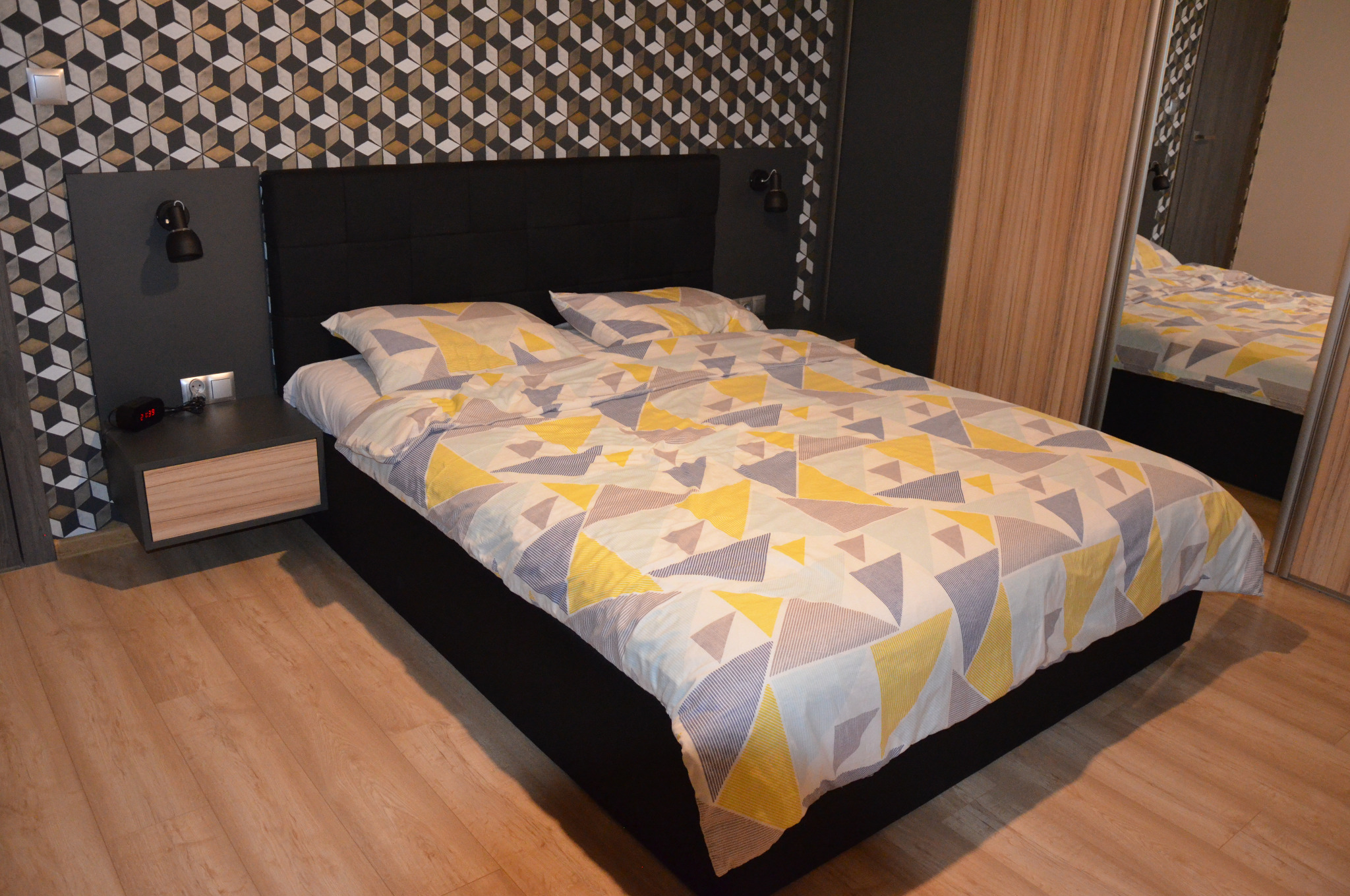 Sypialnia z łóżkiem kontynentalnym, geometryczna tapeta w odcieniach szarości, czerni i złota, drewniana podłoga, szafka nocna z lampką i budzikiem.