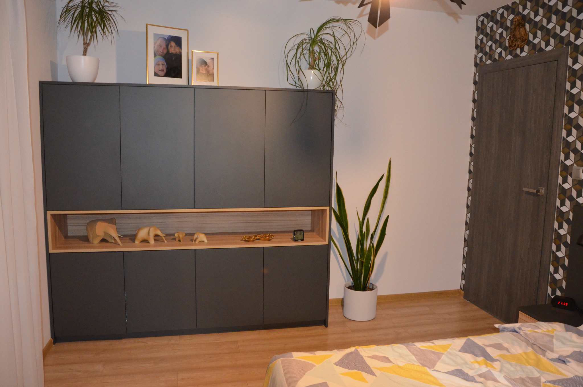 Szara szafa z drewnianymi akcentami w sypialni, na górze rośliny w doniczkach i ramki ze zdjęciami, na półce drewniane figurki zwierząt, obok drzwi z geometrycznym wzorem.