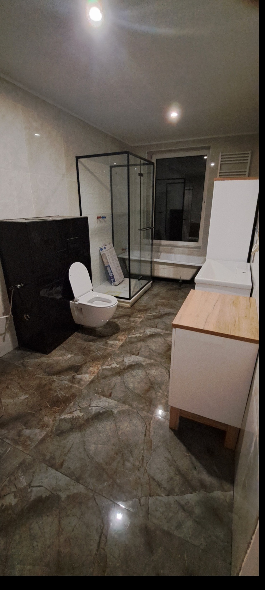 Ukończona łazienka w trakcie montażu: podwieszana toaleta z czarną obudową, kabina prysznicowa z czarnymi ramami i biała szafka z drewnianym blatem.