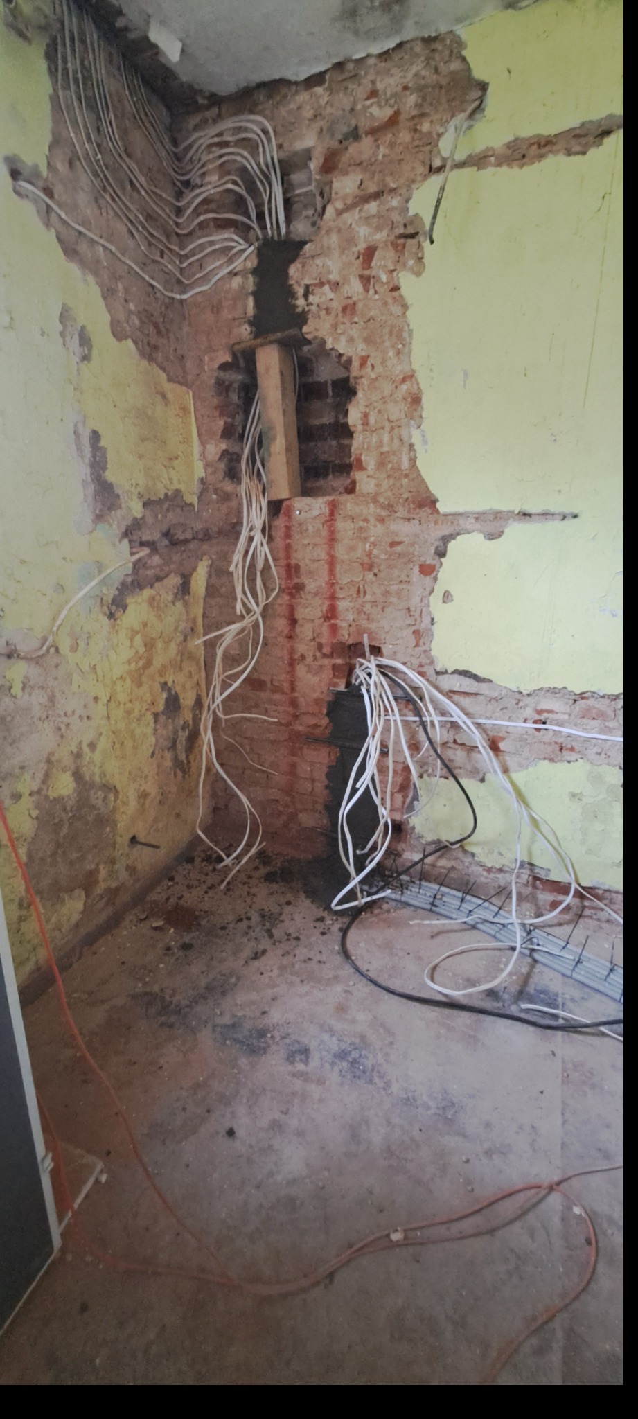 Fragment ściany z odsłoniętą cegłą i przewodami elektrycznymi w trakcie instalacji. Widoczne kable, bruzdy w ścianie i fragment podłogi. Żółte ściany w tle.