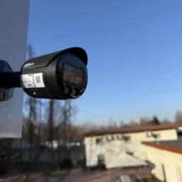 Czarna kamera monitoringu Dahua zamontowana na białym słupku, widok z dołu na tle błękitnego nieba i rozmytego budynku z drzewami w tle.