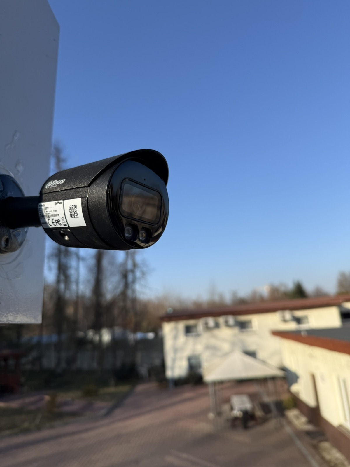 Czarna kamera monitoringu Dahua zamontowana na białym słupku, widok z dołu na tle błękitnego nieba i rozmytego budynku z drzewami w tle.