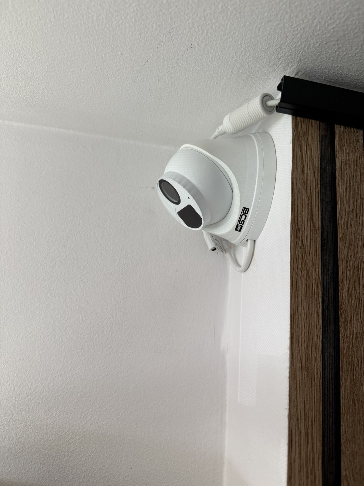 Kamera kopułkowa BCS zamontowana w rogu pomieszczenia, widoczne przewody i element dekoracyjny z drewnianymi lamelami.